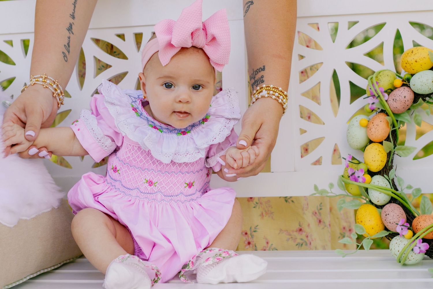 Baby girl in pink smocked romper sitting on white bench with colorful Easter egg decorations nearby.