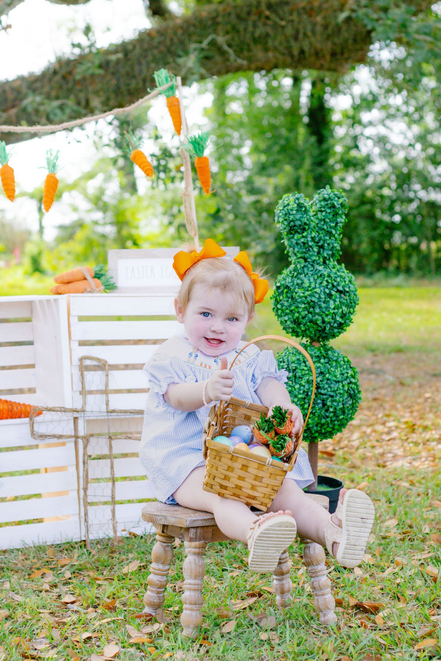 Toddler girl with carrot headband holds Easter basket while sitting near bunny topiary outdoors.