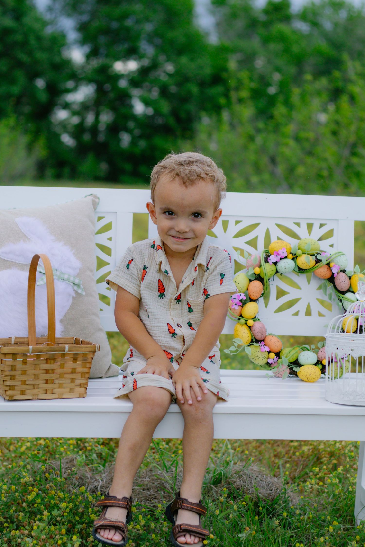 Adorable young boy in carrot-print romper sits on white bench beside Easter basket and floral wreath in a garden setting.