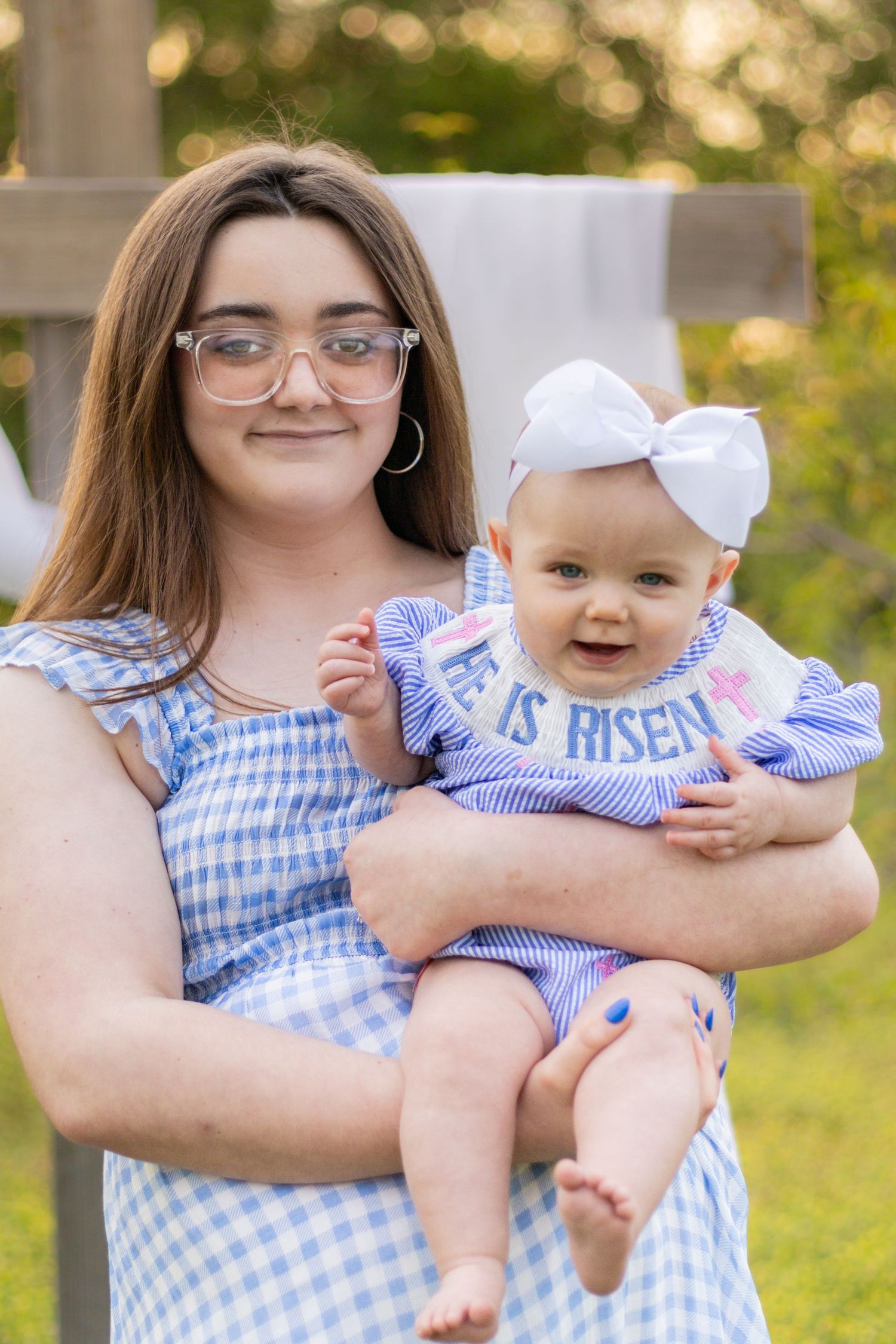 Young mother wearing glasses holding smiling baby in white bow and He Is Risen bib in outdoor setting.