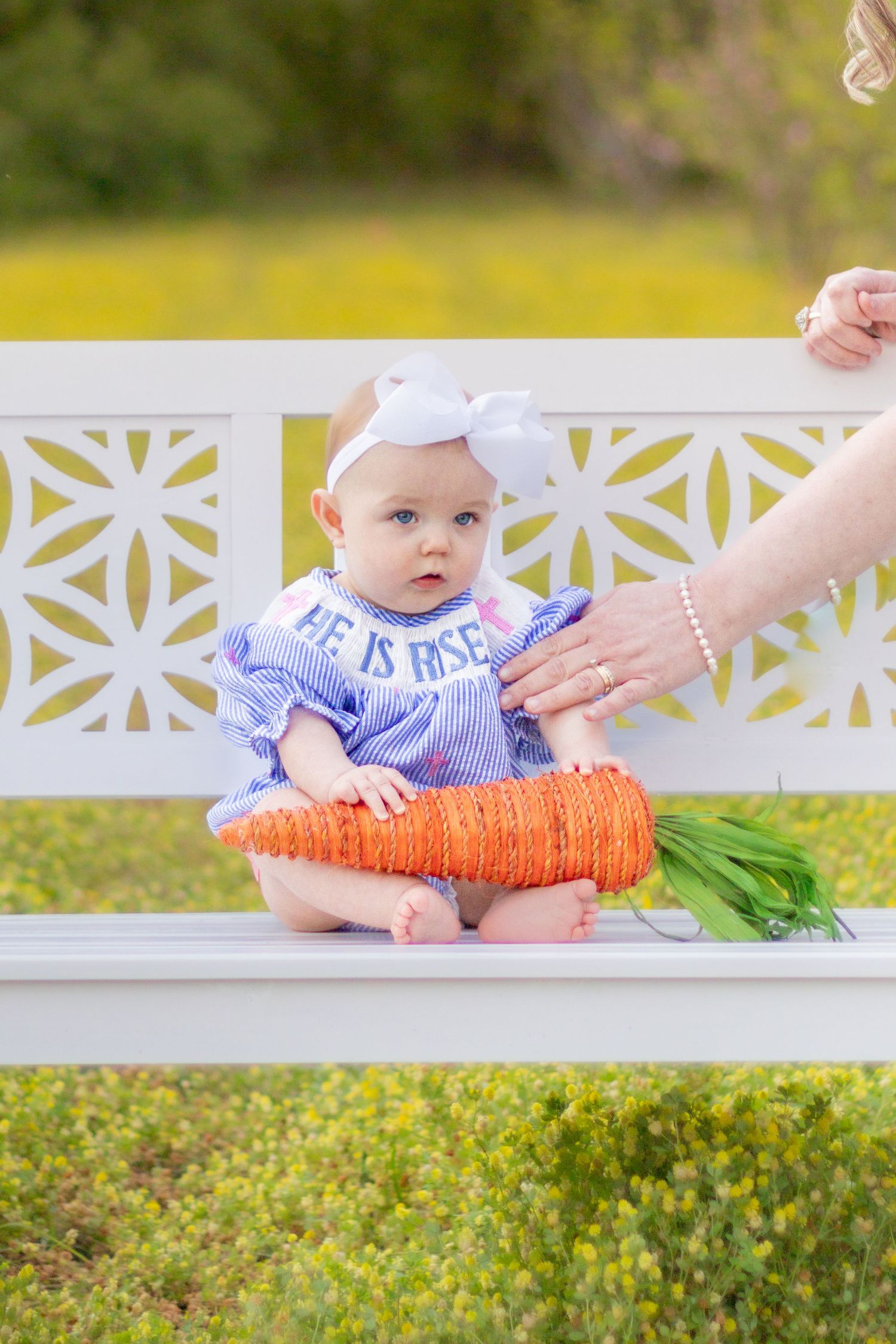Baby in blue striped He Is Risen dress holding decorative carrot on white bench outdoors.