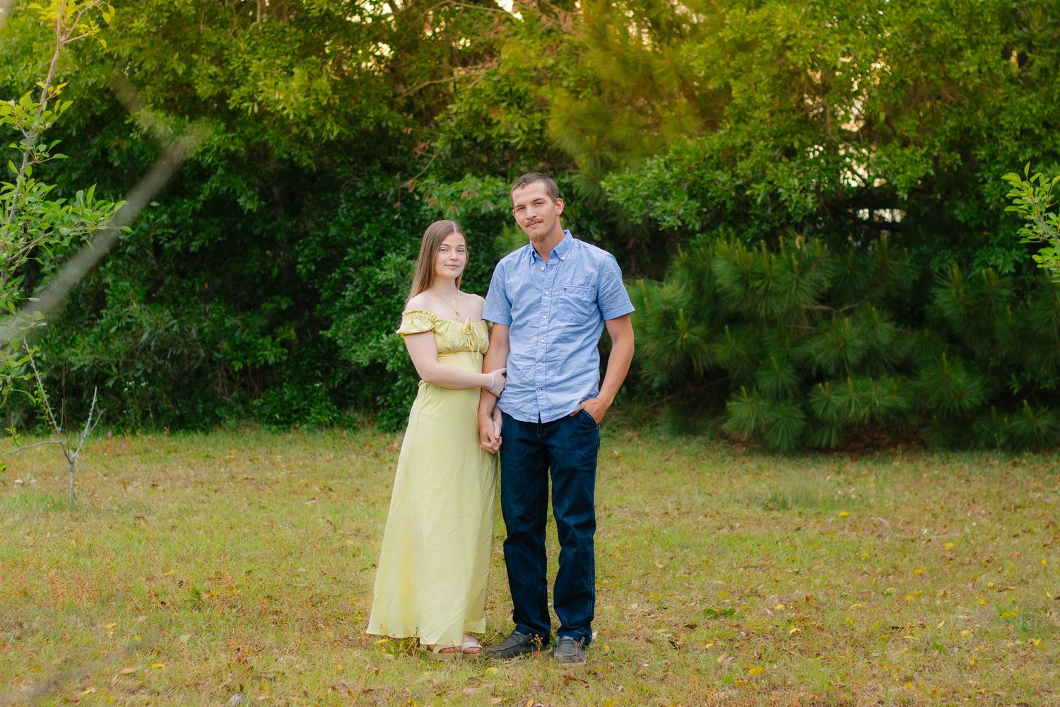 Smiling couple standing together in grassy field with green trees in the background.
