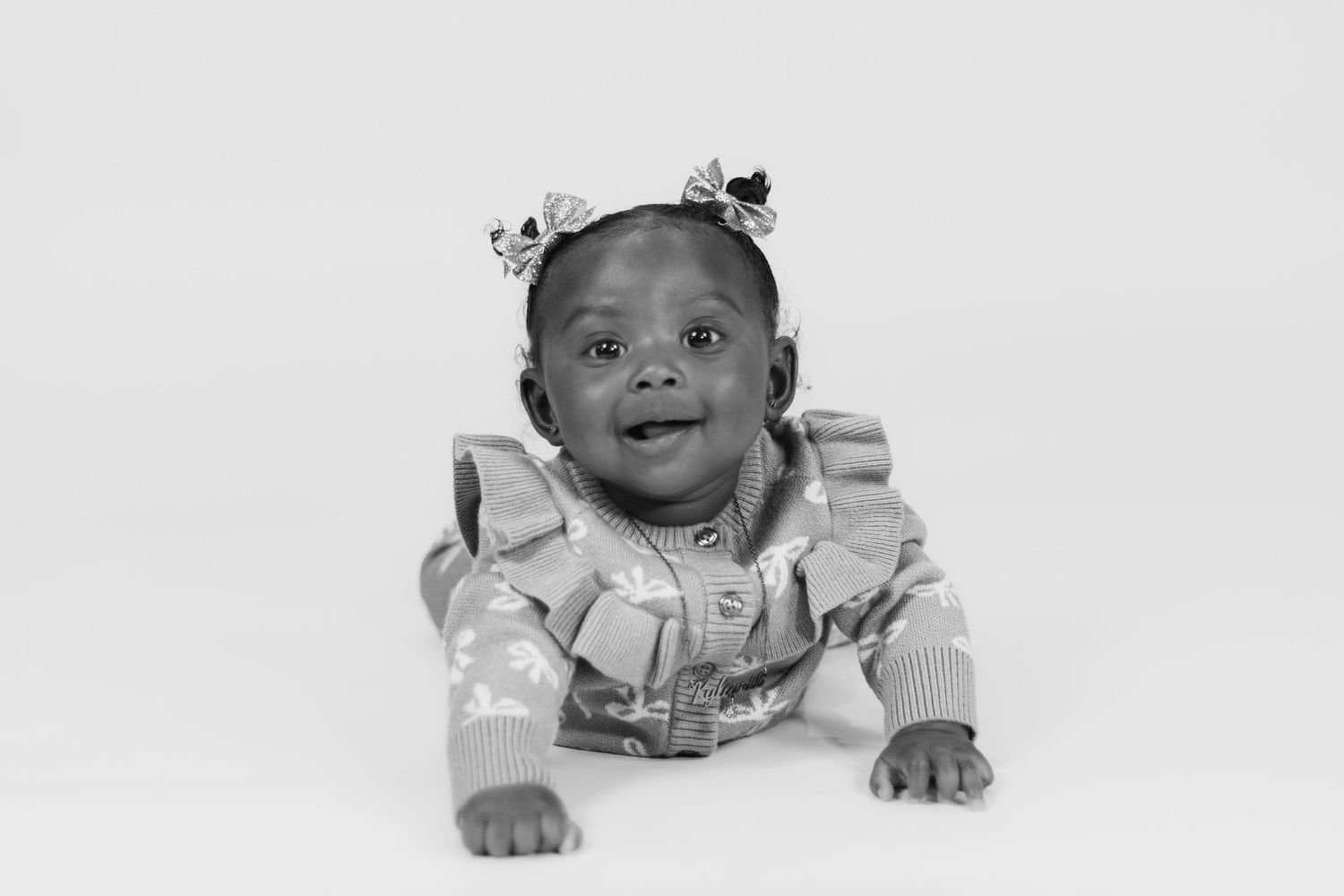 Adorable baby girl in a ruffled sweater and floral headband smiling while doing tummy time, black and white photo.