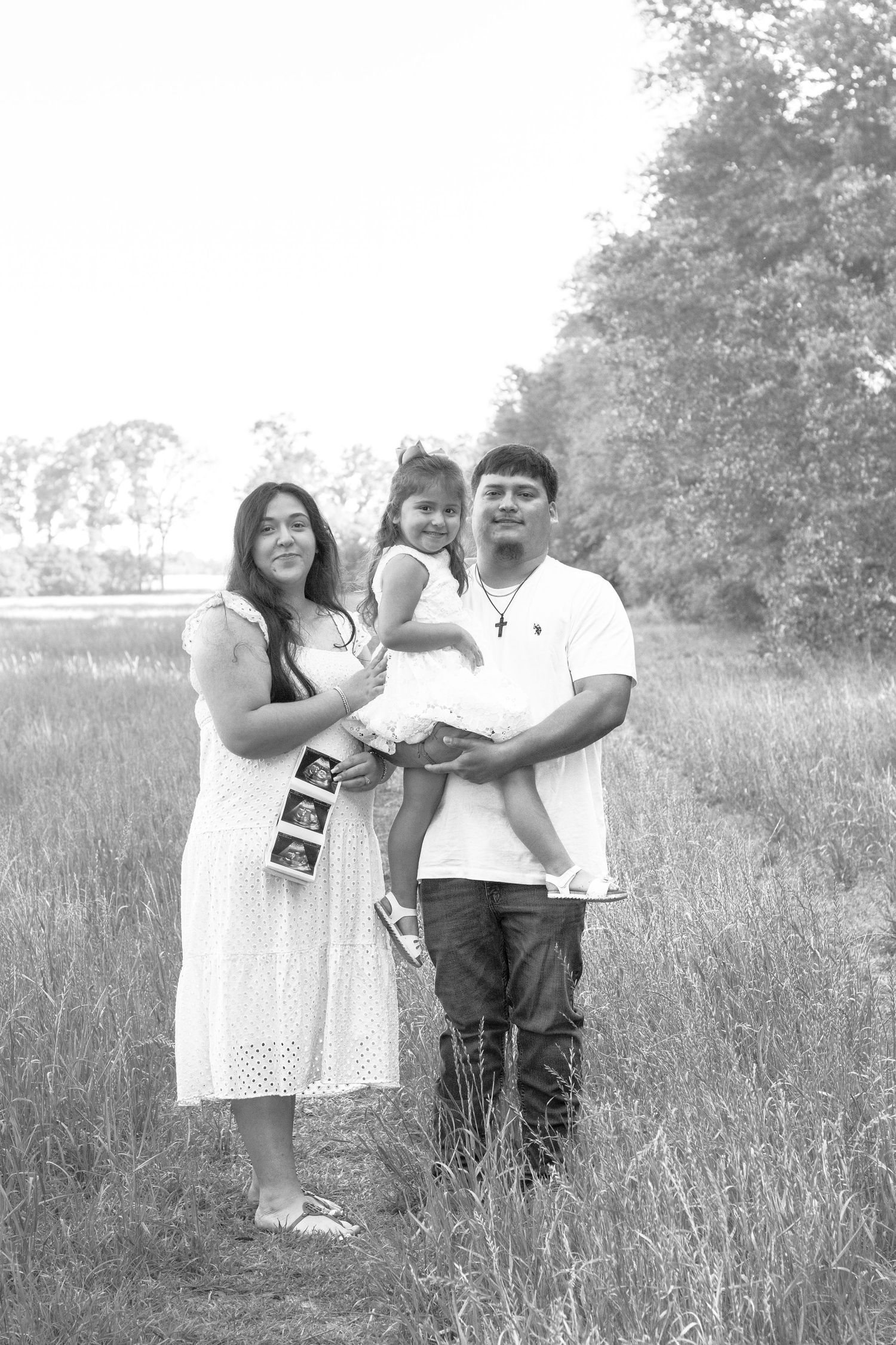 Black and white family maternity photo of couple holding toddler girl in field, displaying ultrasound pictures outdoors.