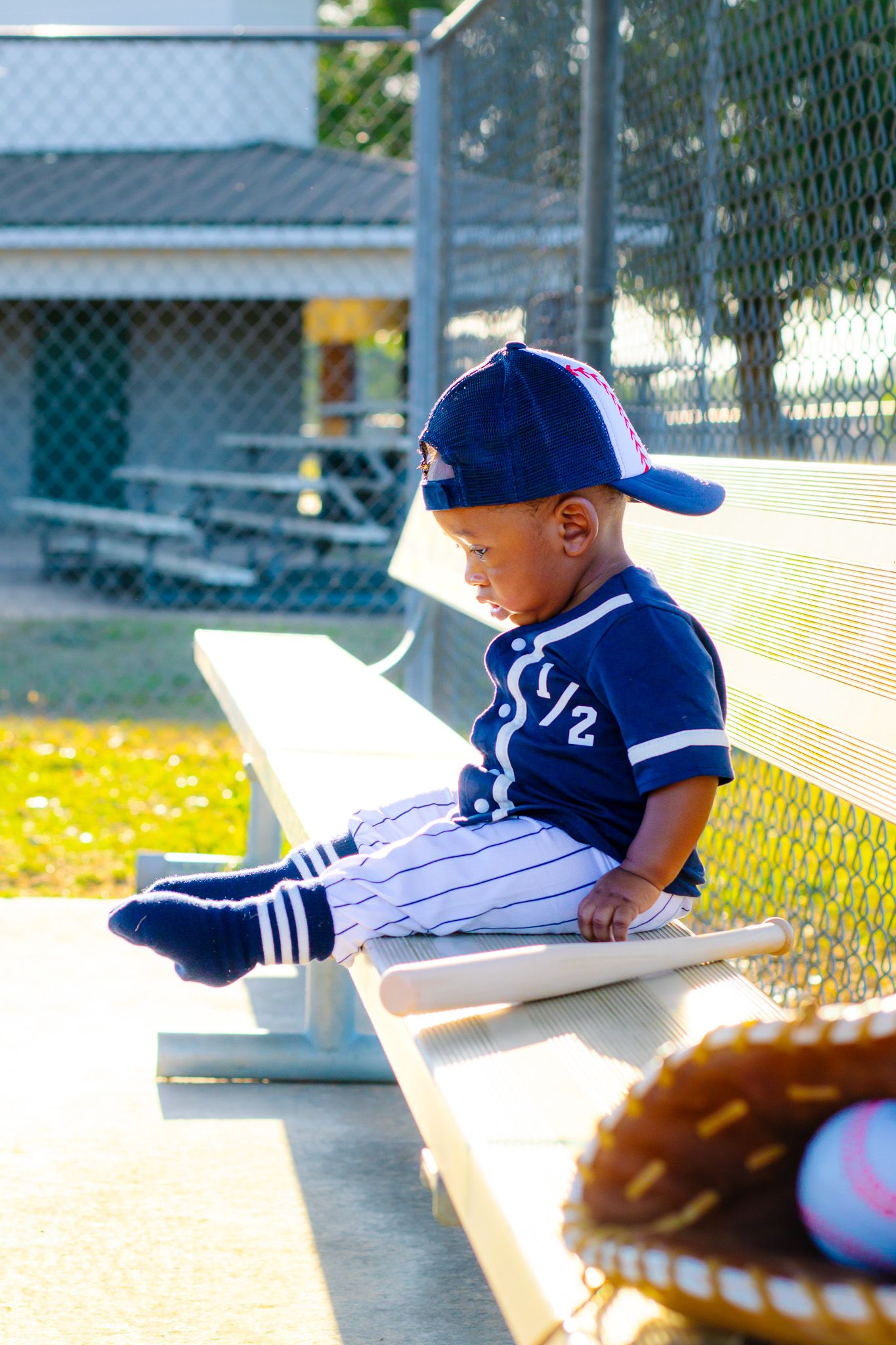 Young toddler in blue baseball uniform sitting on dugout bench with bat and glove at youth baseball field.