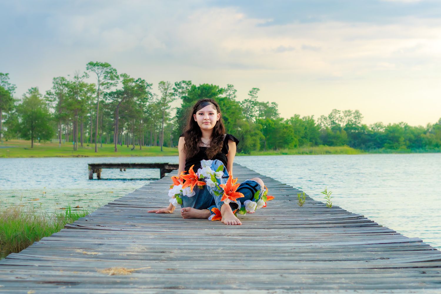 Young girl sitting cross-legged on a wooden dock holding colorful flowers, with a serene lake and trees in the background.
