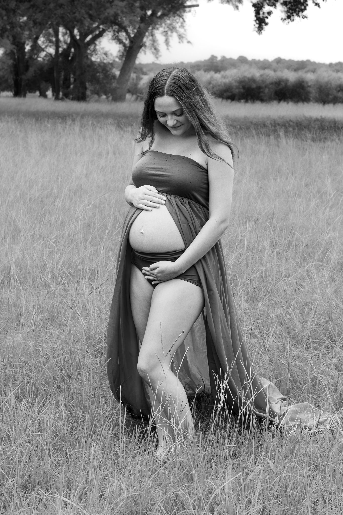 Black and white photo of a pregnant woman in a flowing dress cradling her baby bump in a grassy field.