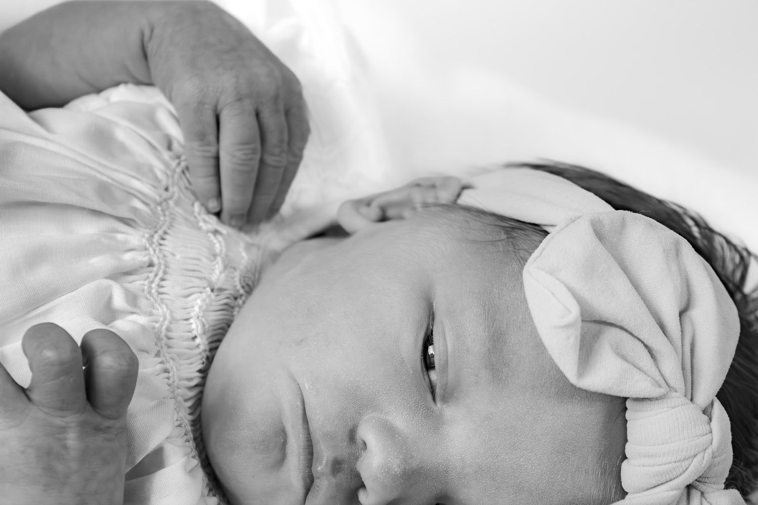 Black and white newborn baby girl wearing a white bow headband, lying peacefully in soft fabric.