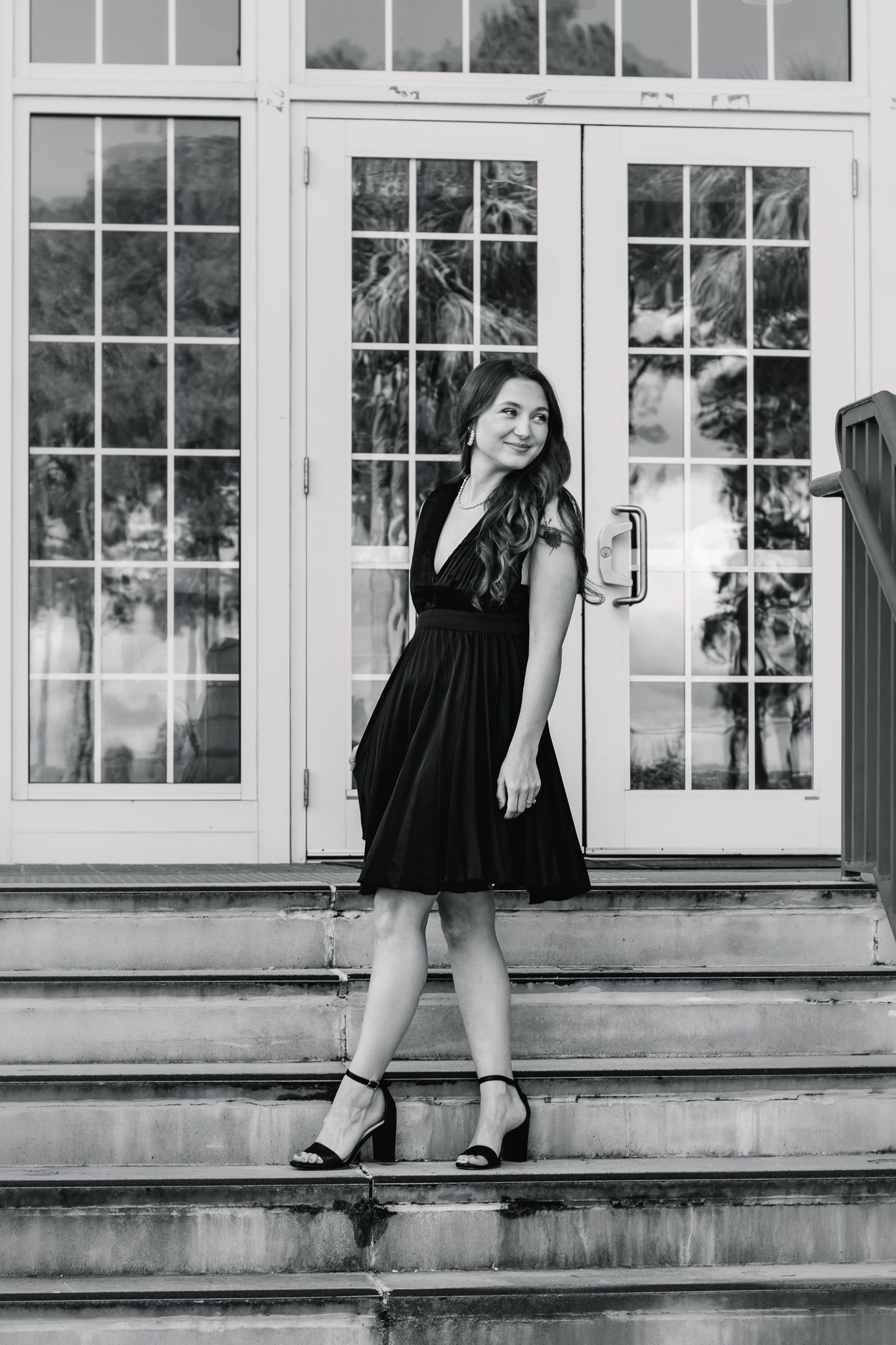 Black and white photo of a young woman in a black dress and heels posing on stone steps outside elegant French doors.