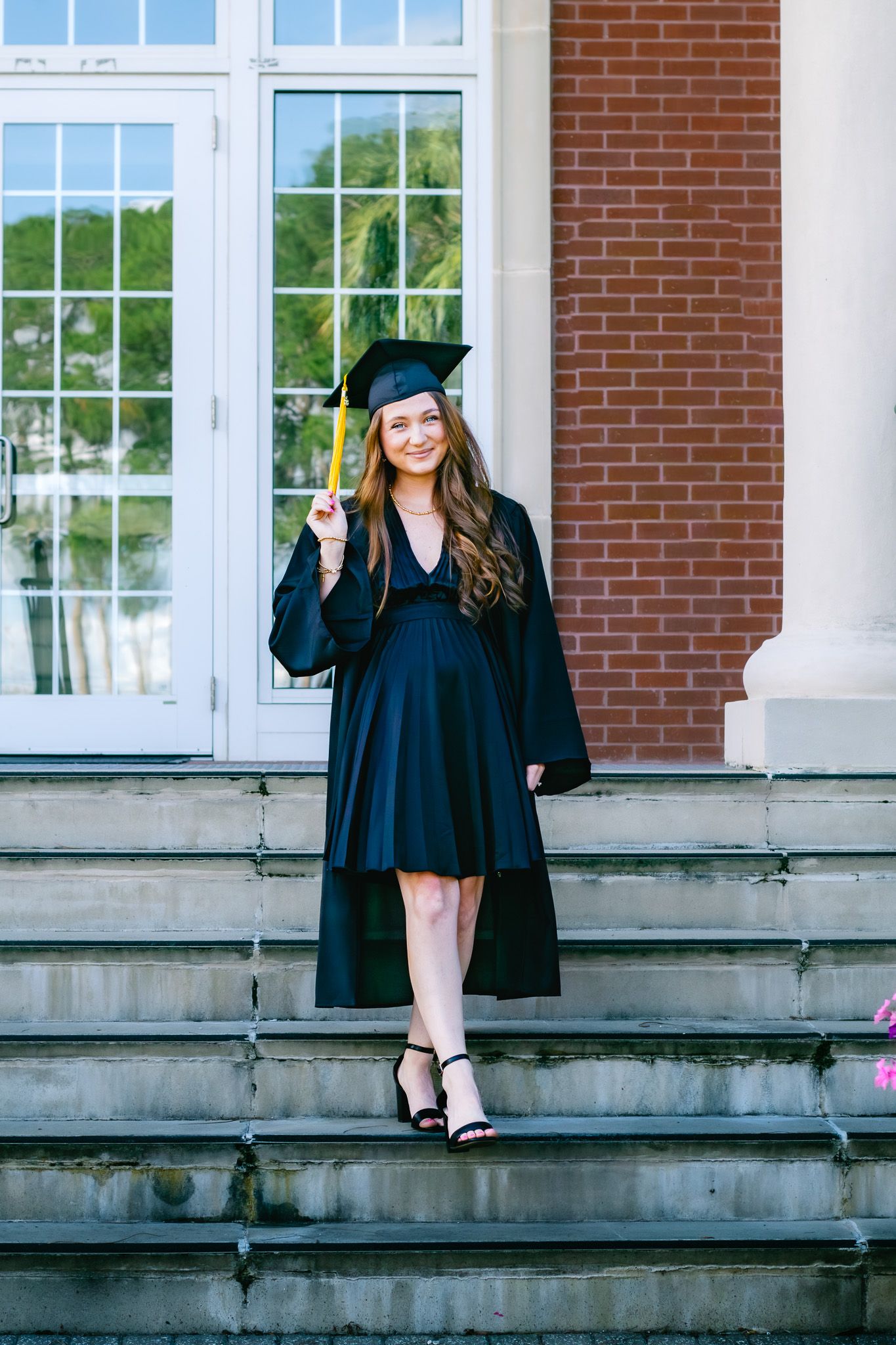 A smiling young woman in black graduation cap and gown poses proudly on university steps.