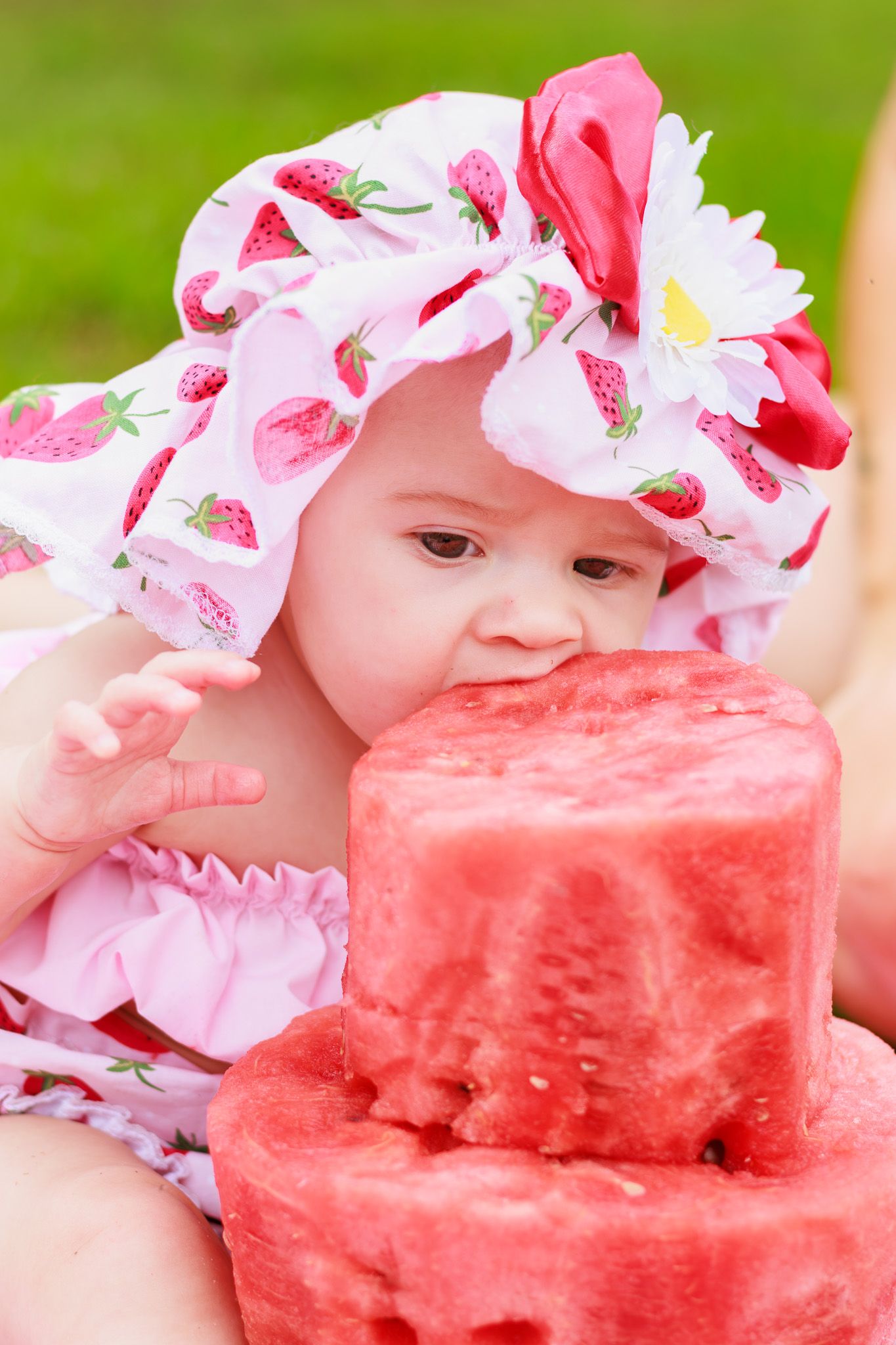 Close-up of a cute baby in a floral strawberry hat eagerly eating a big chunk of fresh watermelon outside.