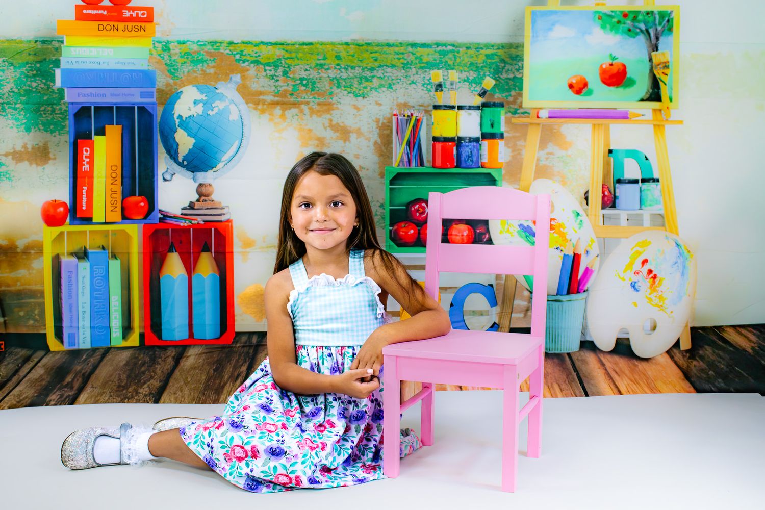 Young girl in floral dress sits beside pink chair with colorful school-themed backdrop featuring books and globe.