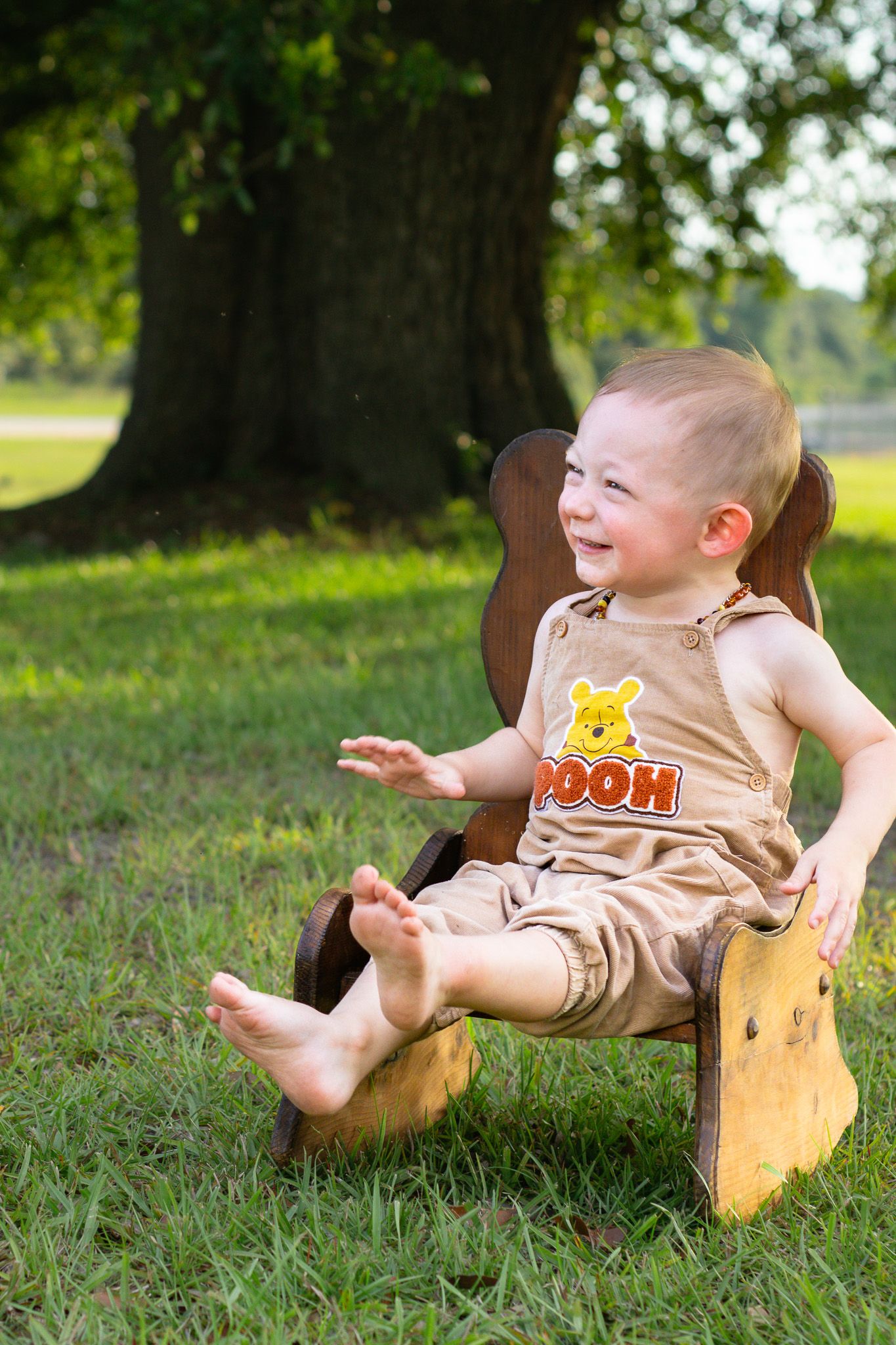 Smiling baby boy in Winnie the Pooh overalls sitting in a wooden chair on green grass near a large tree.