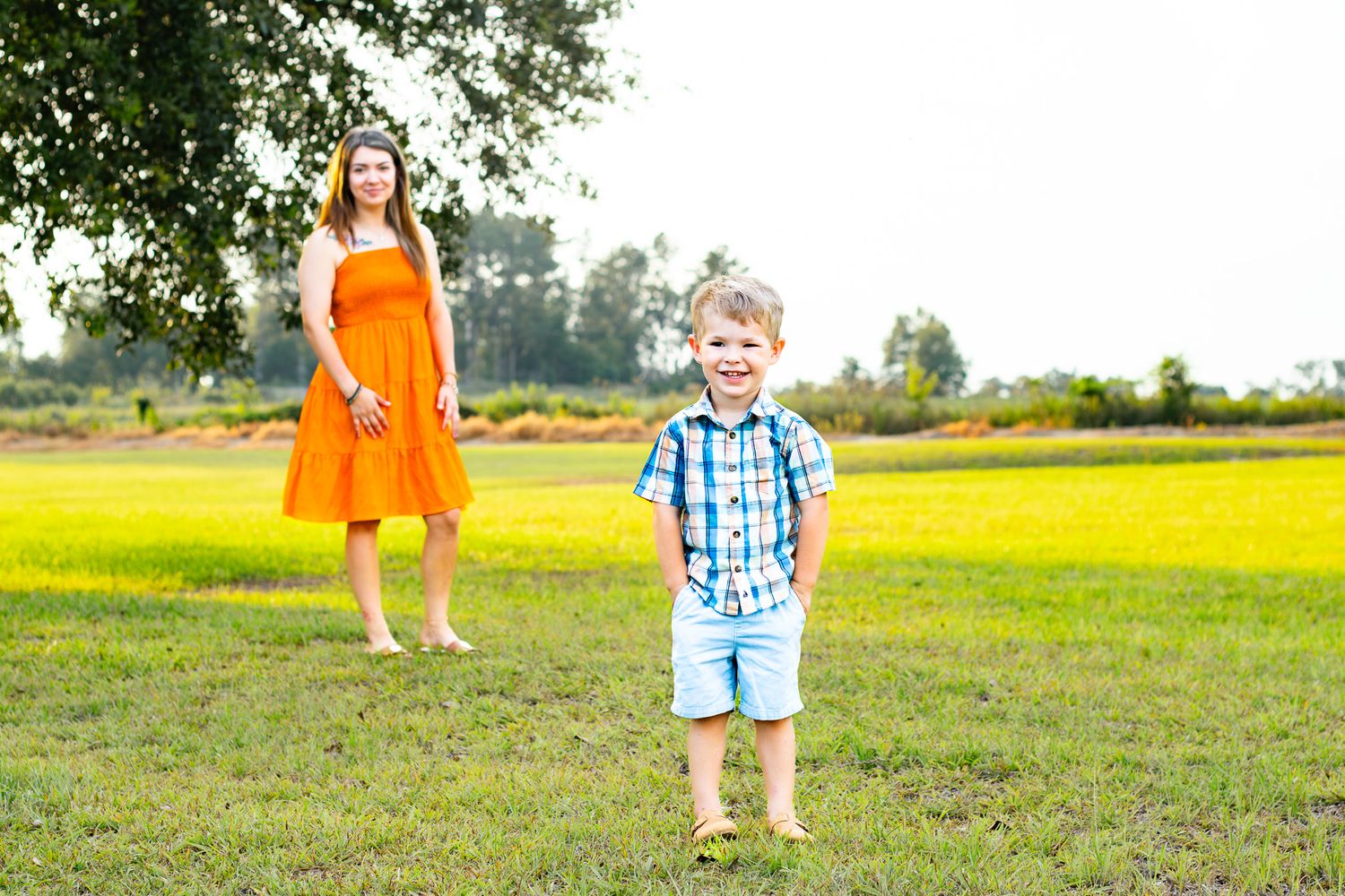 A young boy in a plaid shirt smiles on green grass while a woman in an orange dress stands behind him outdoors.