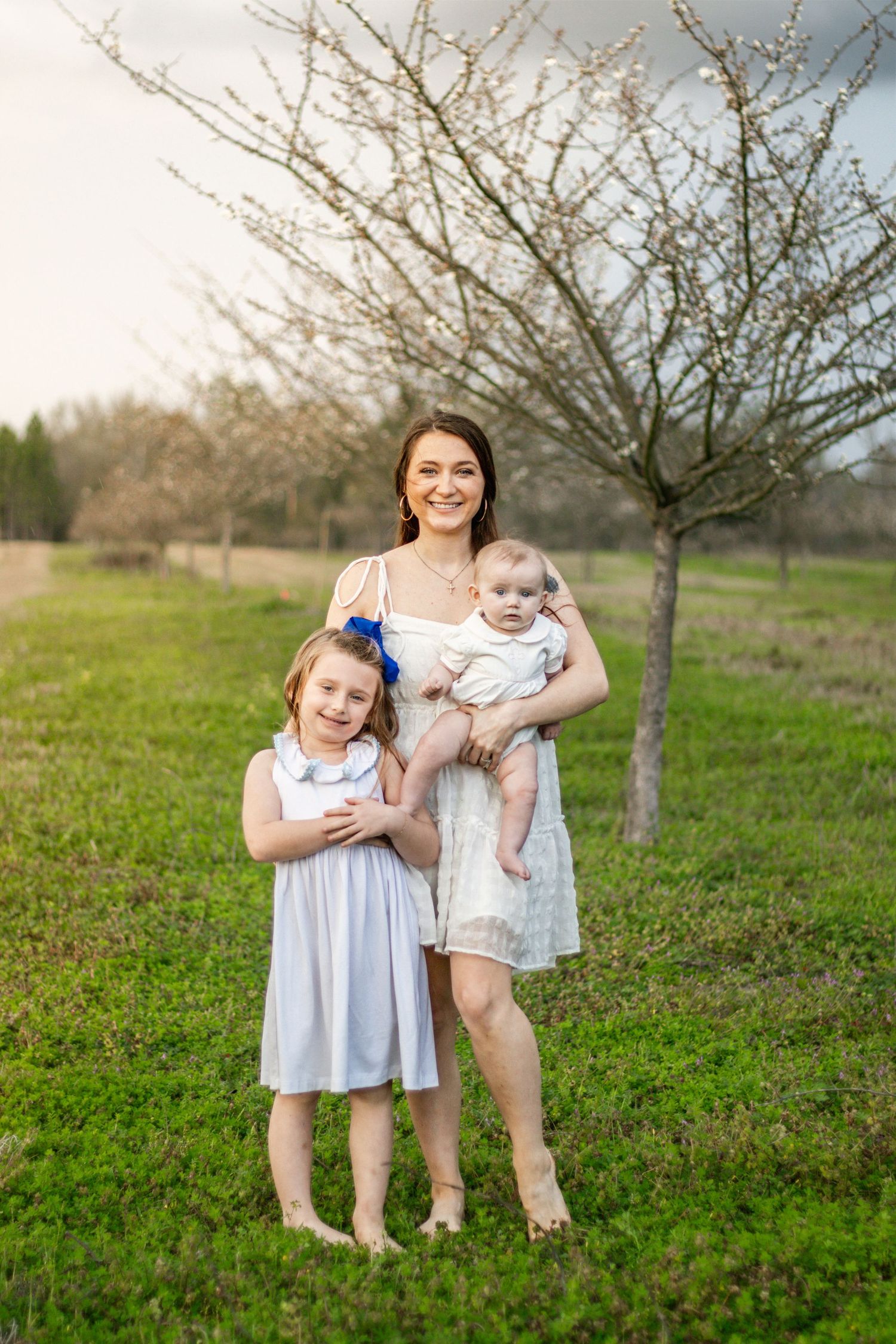 Mother holding baby standing with daughter near blooming tree outdoors.