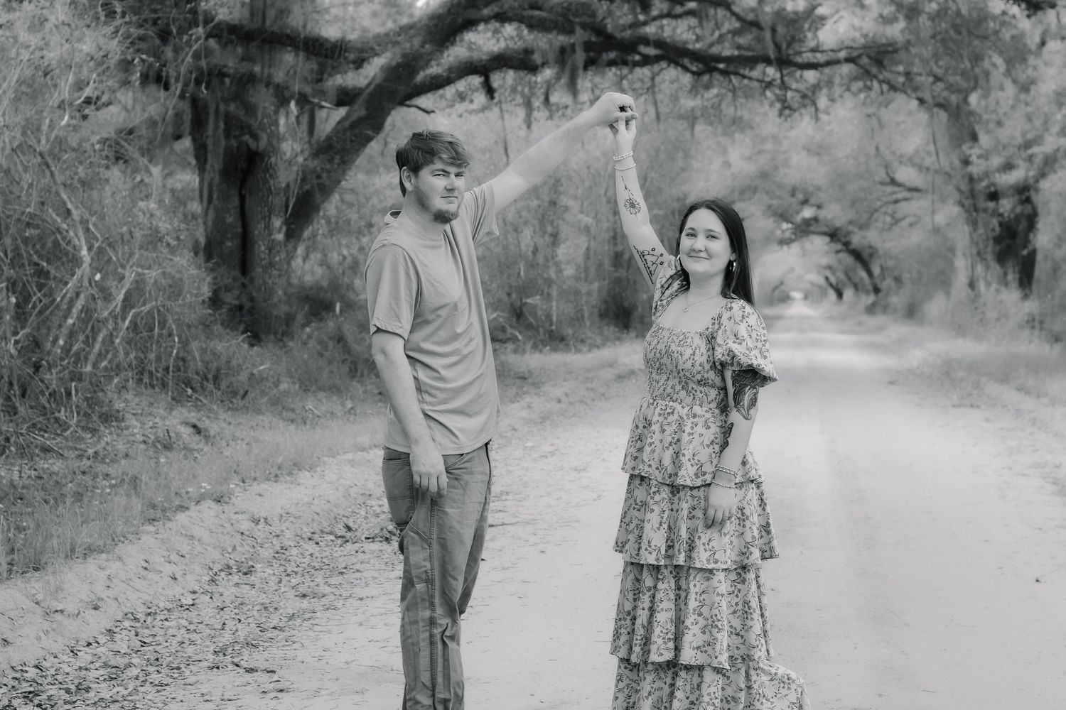 Black and white photo of a couple dancing on a tree-lined dirt road, man spinning woman in floral dress.