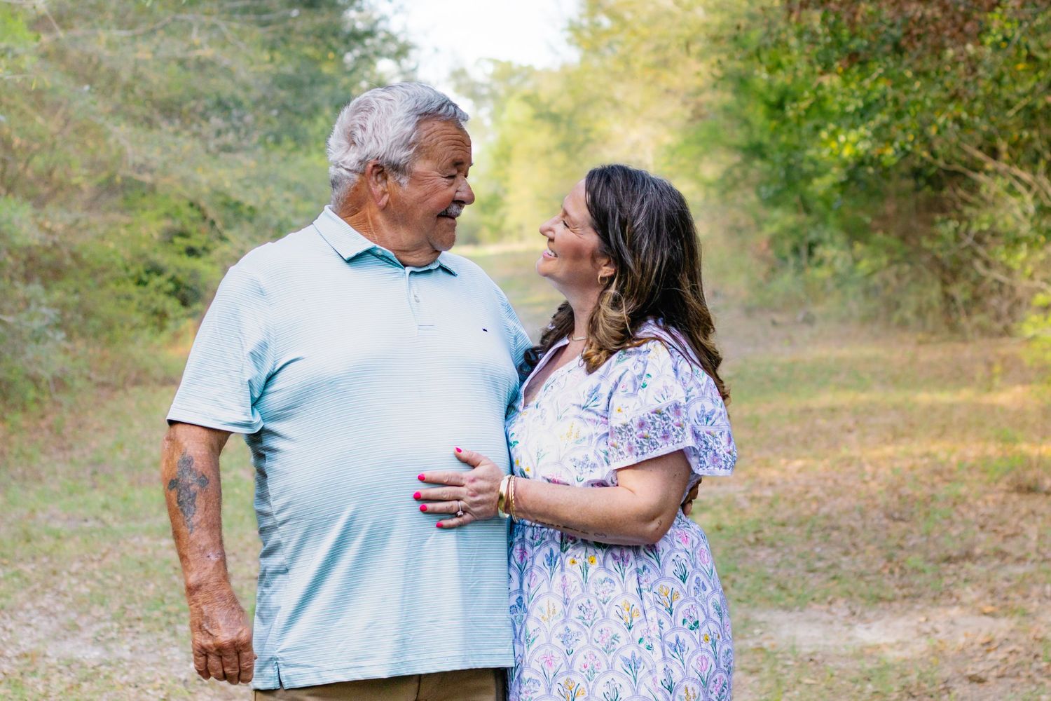 Older couple sharing a tender moment on a wooded path, man in blue polo and woman in floral dress.