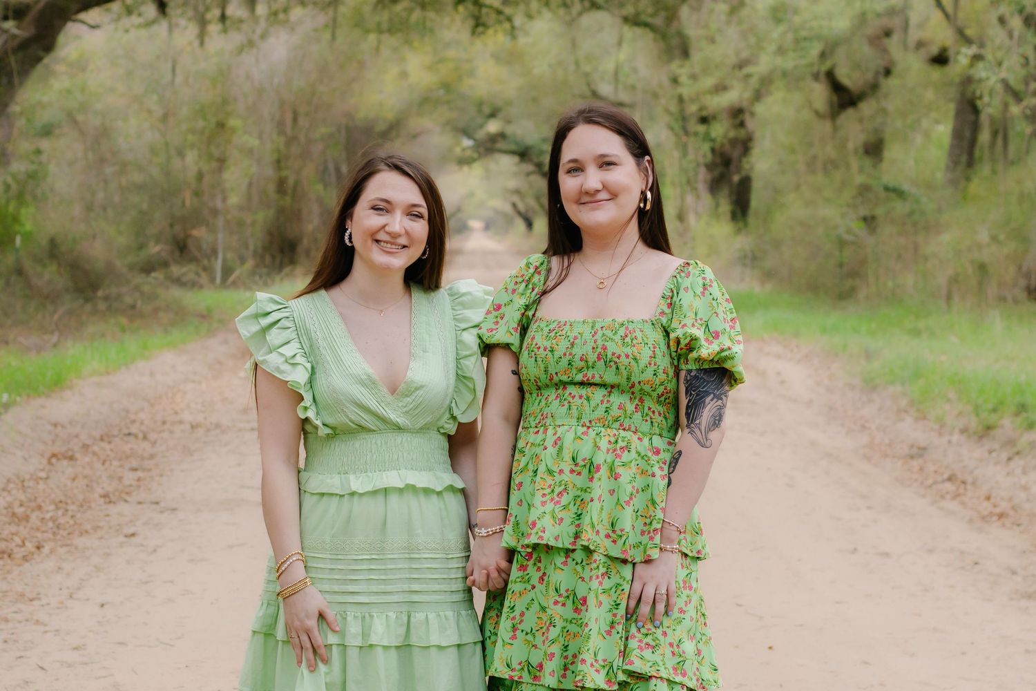 Two smiling women in green dresses posing together on a rustic dirt road surrounded by trees.
