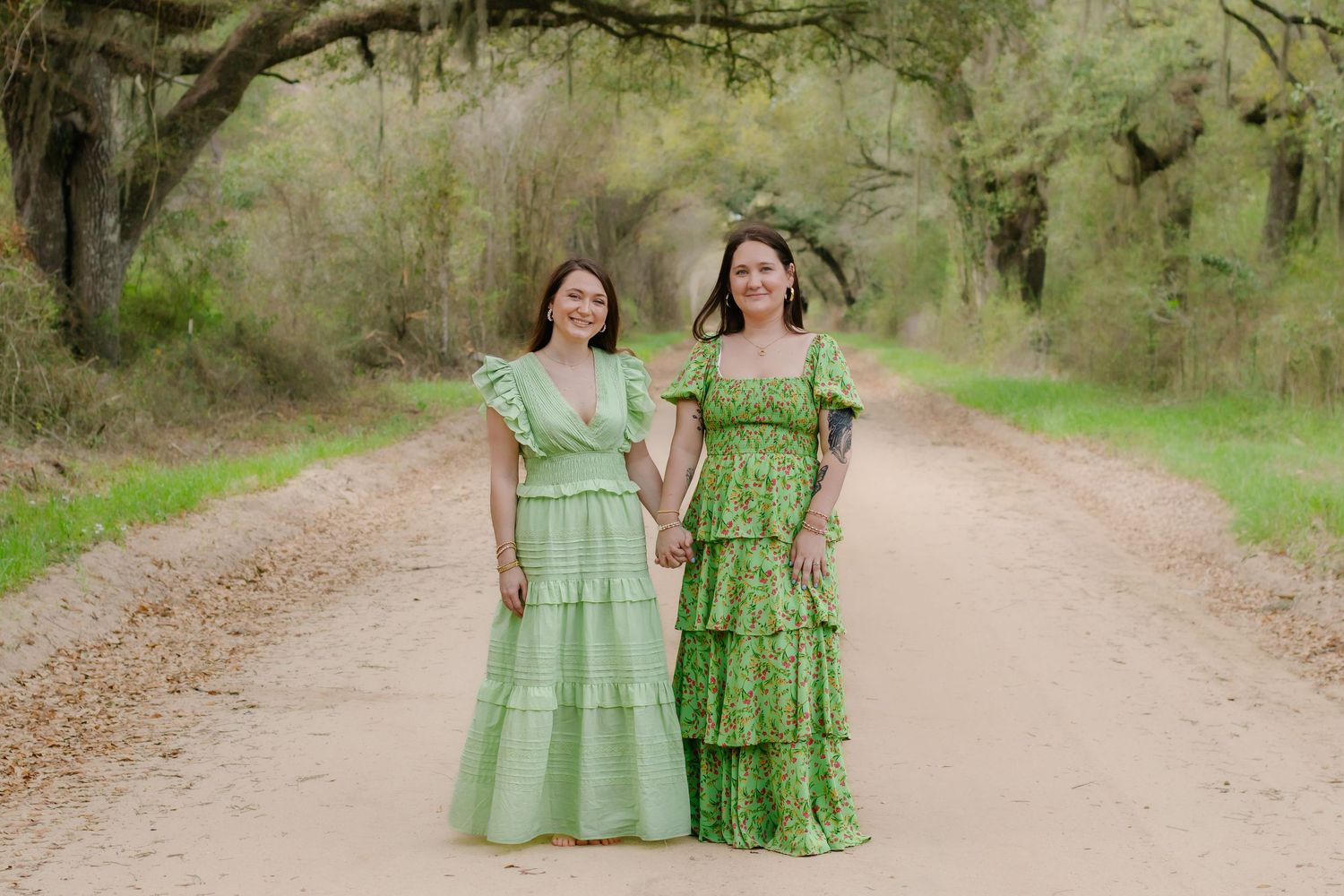 Two women in green dresses hold hands on a scenic dirt road.