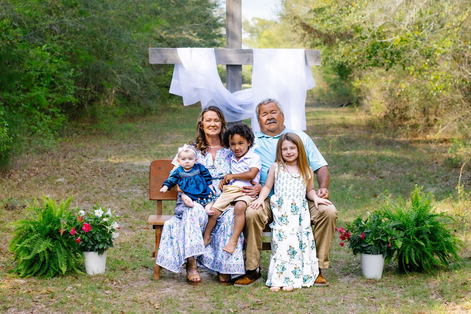 Family of five with children posing on wooden bench before a large cross with white draping outdoors.