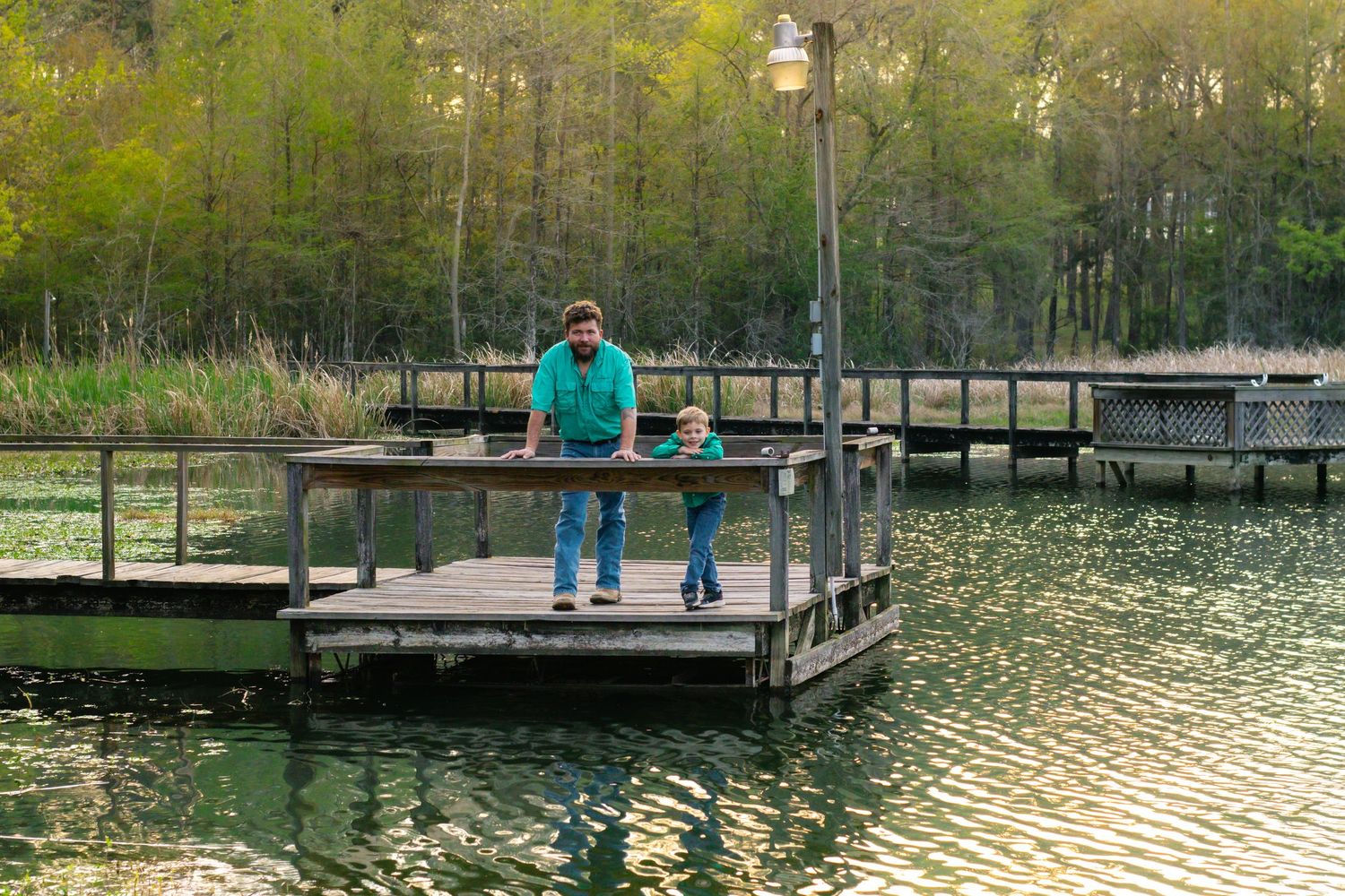 Father and young son stand together on a wooden dock surrounded by a calm green lake.