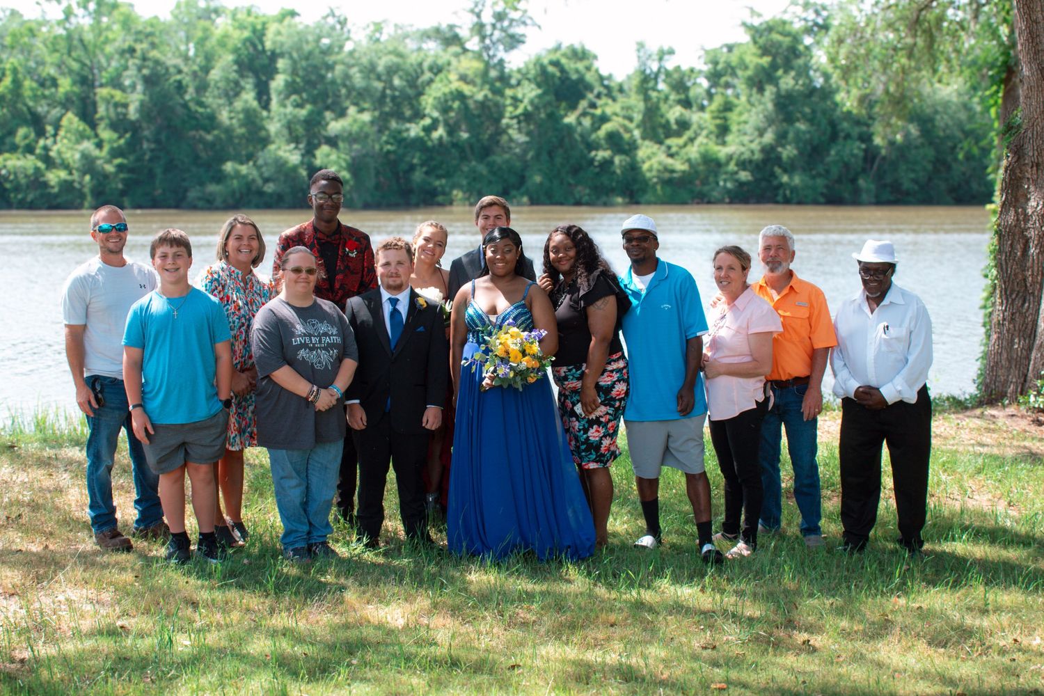 A large group of family and friends gathers outdoors near a river to celebrate a prom occasion.
