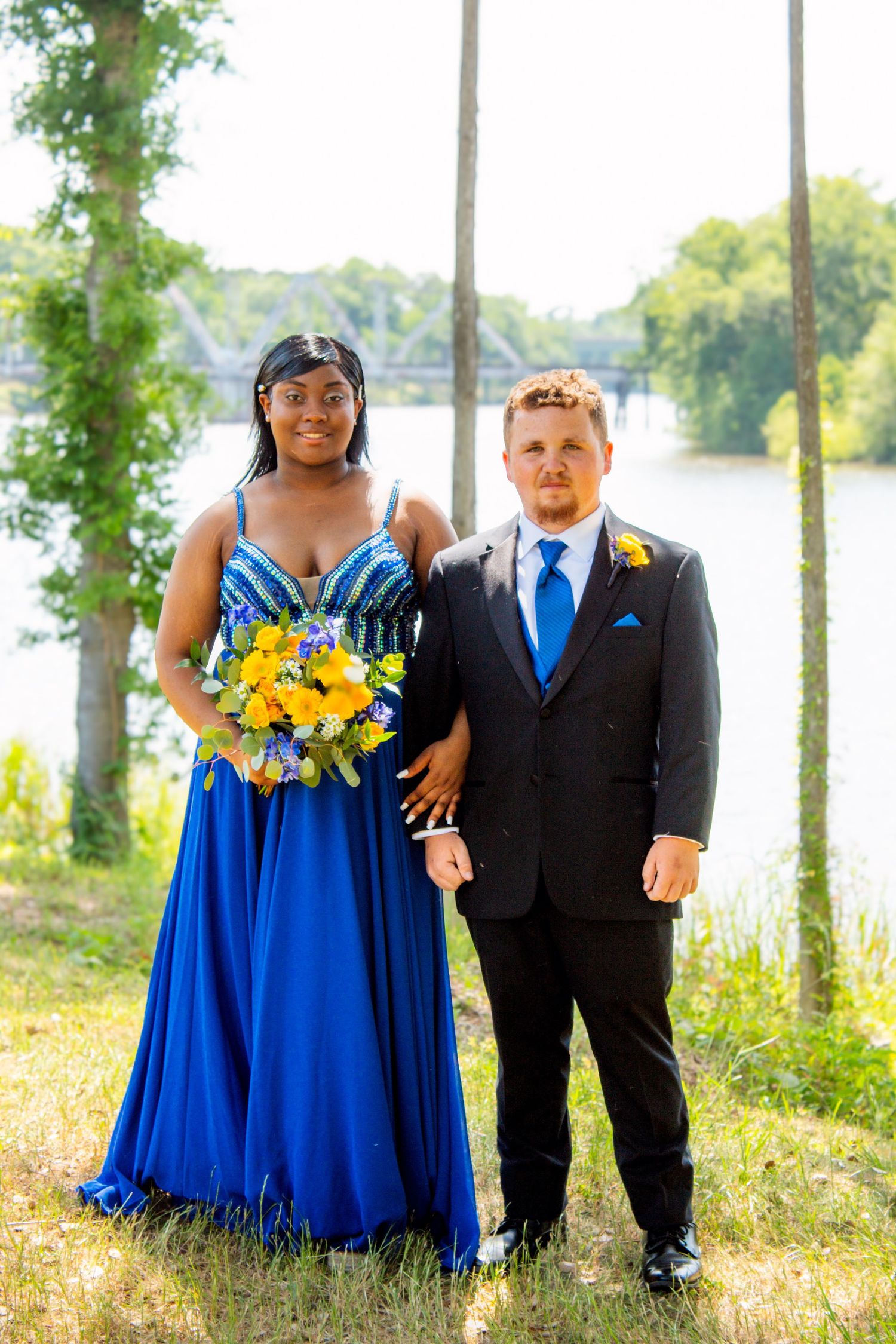 A prom couple stands by a river, woman in royal blue gown holding yellow floral bouquet.