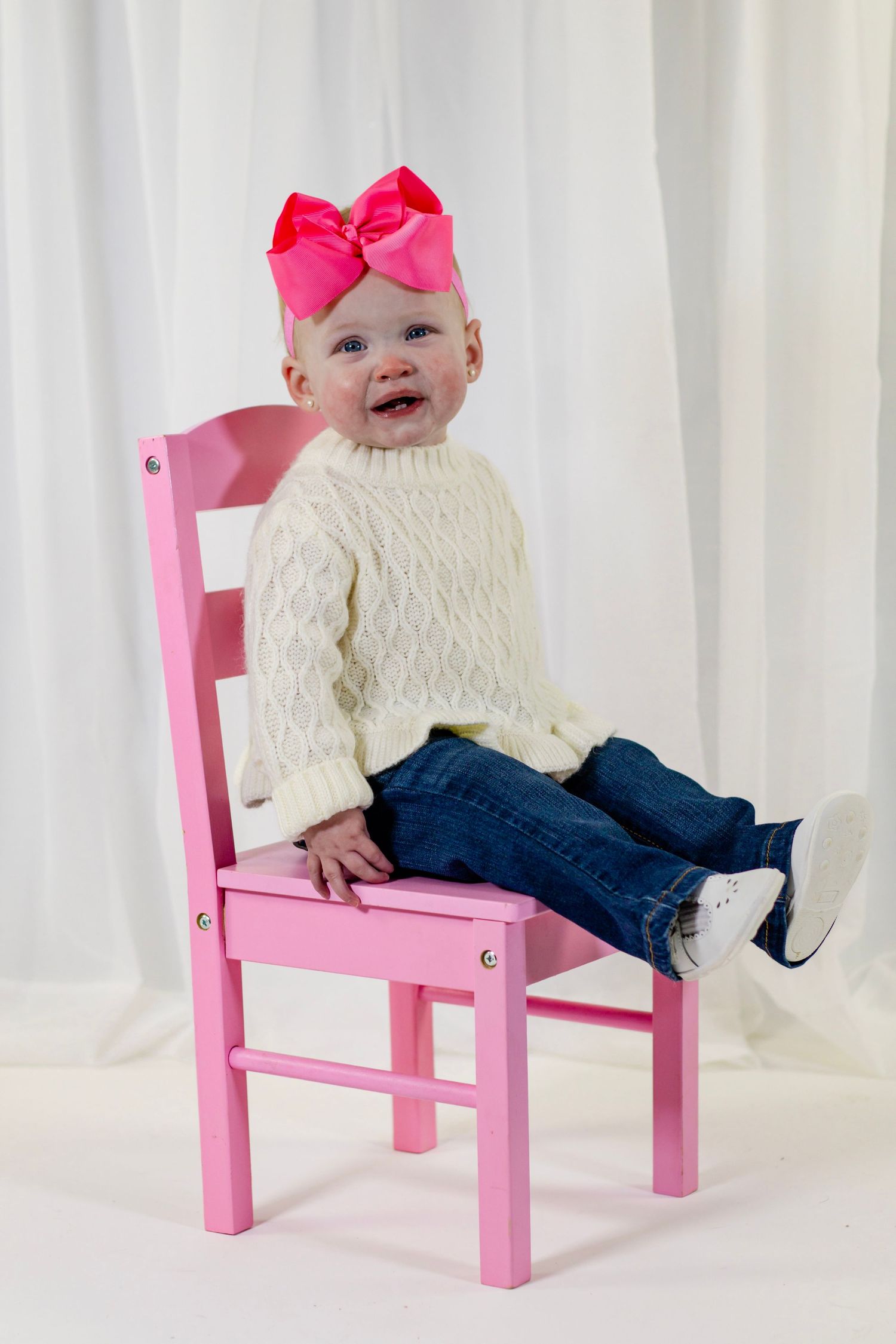Smiling baby girl in white cable-knit sweater and pink bow sitting on a pink wooden chair.