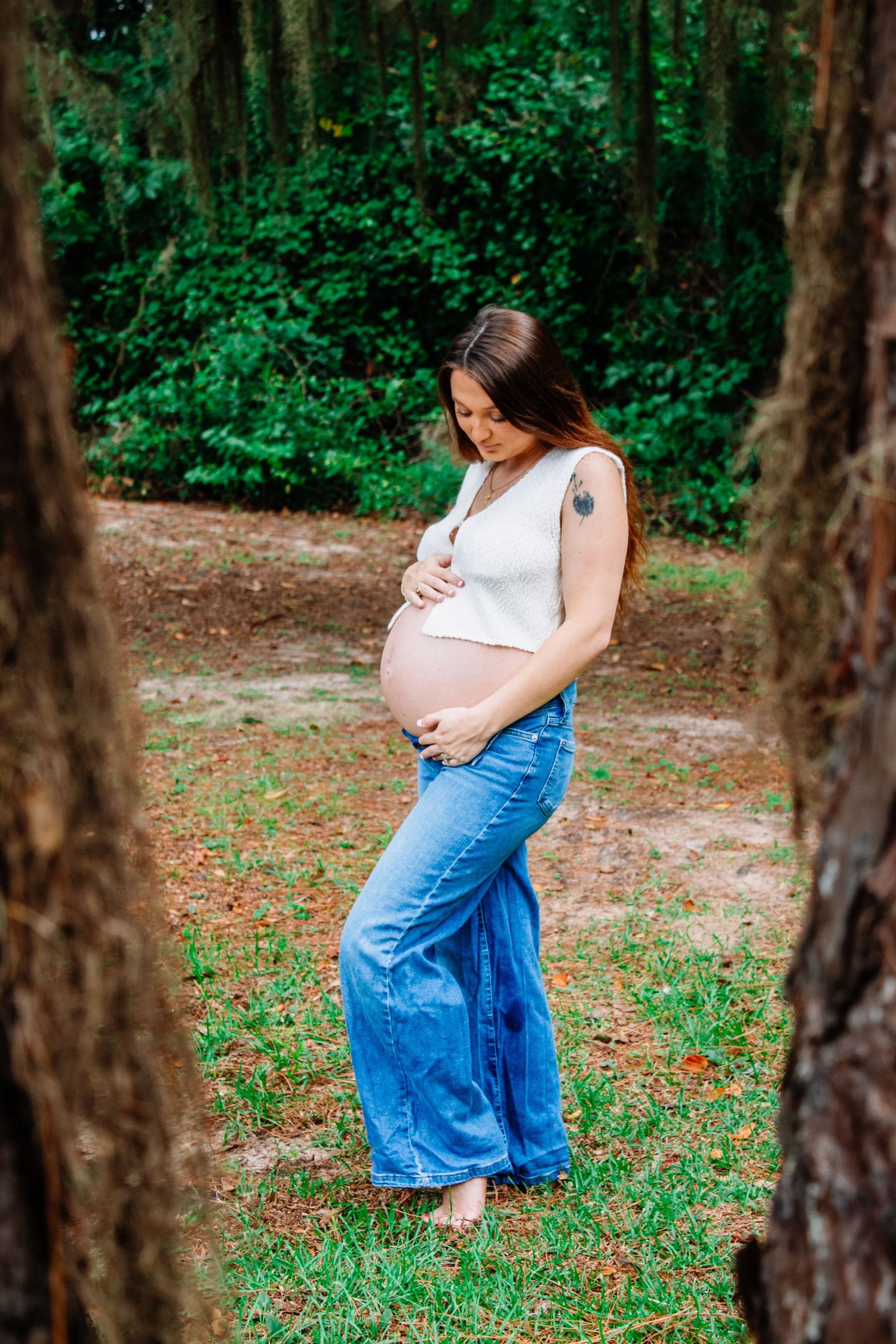 Pregnant woman in white top and blue jeans cradling baby bump while standing between trees in a park.