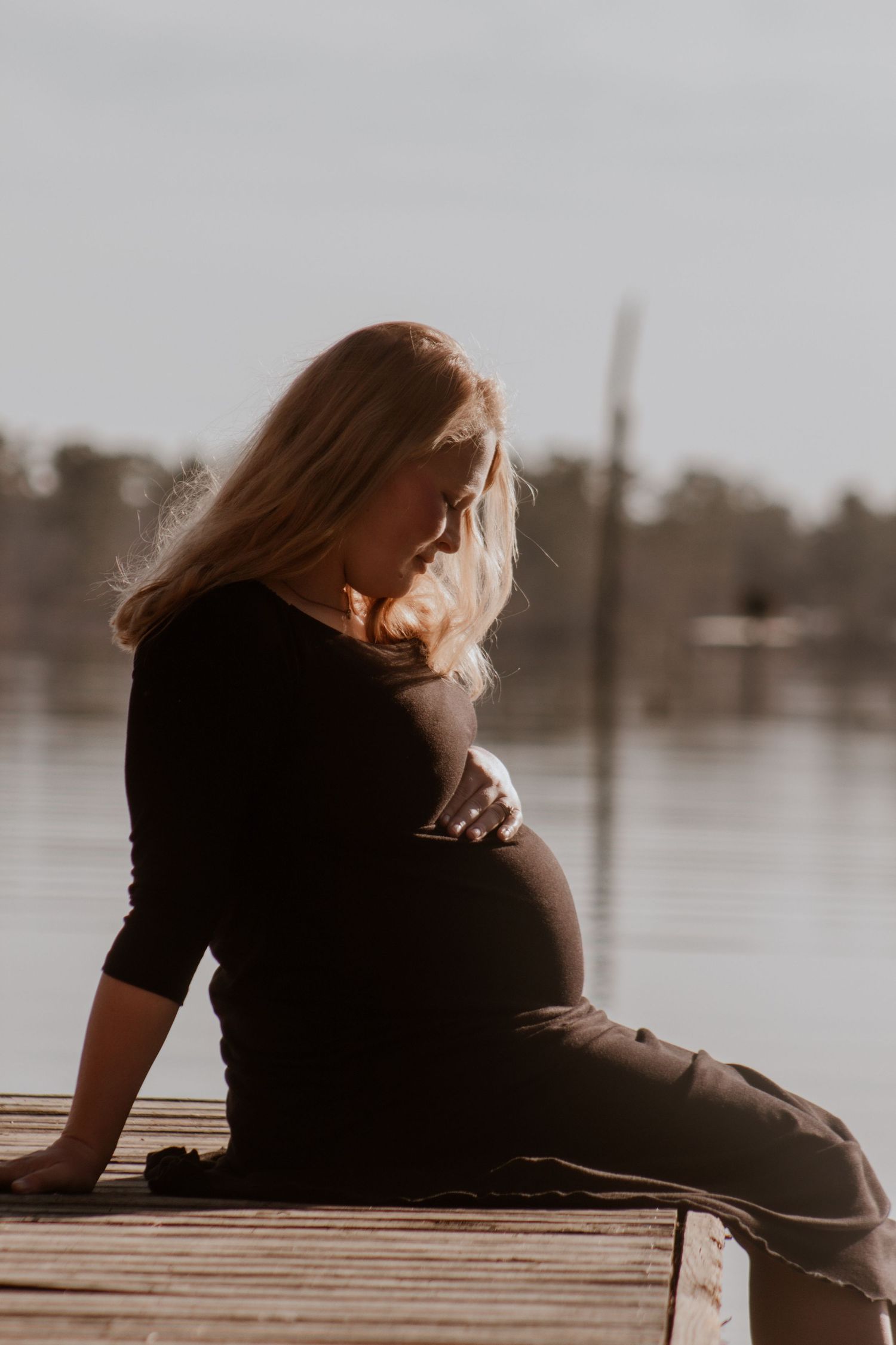 Pregnant woman in black dress sitting on wooden dock by lake looking down at baby bump in soft light.