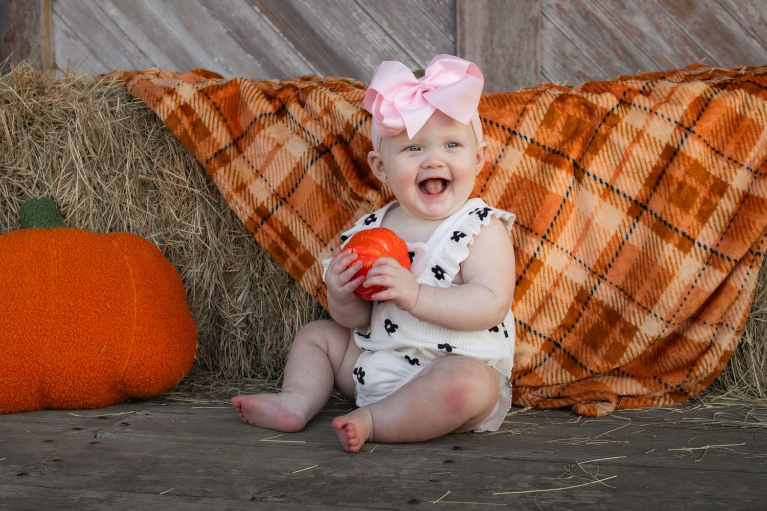 Happy baby girl with pink bow headband laughing while holding small pumpkin in fall setting.
