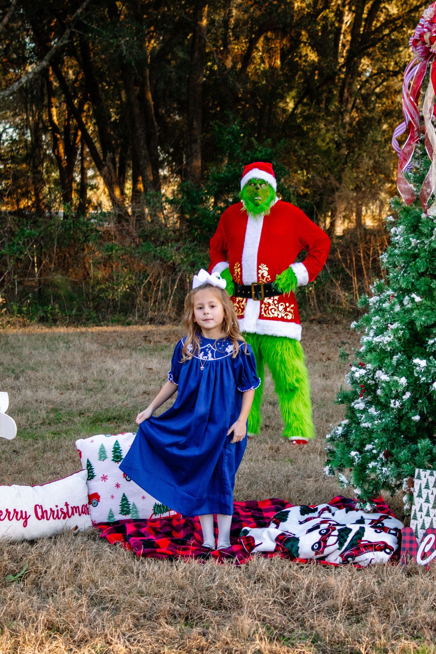A young girl in a blue dress poses with the Grinch in a Santa suit beside a Christmas tree outdoors.