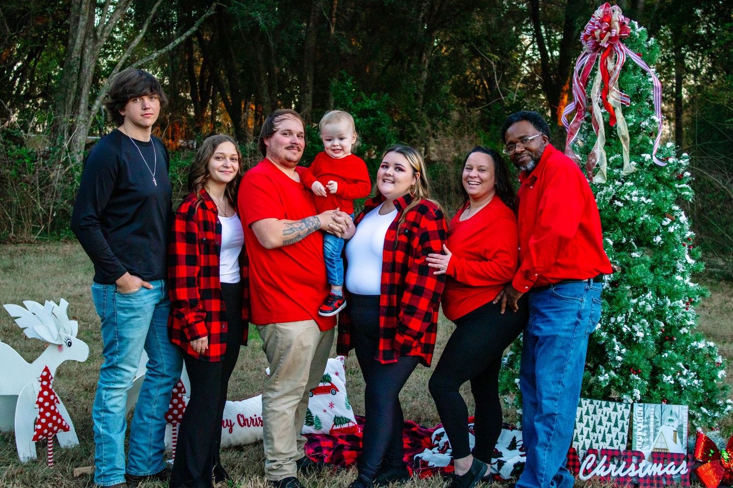 A family of seven in red and plaid outfits smiles together beside a decorated Christmas tree outdoors.