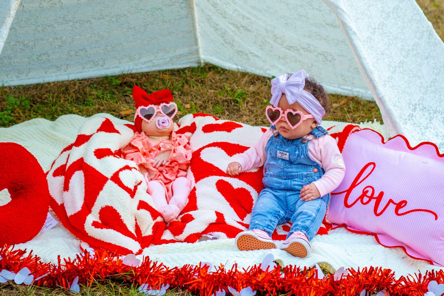 Two babies in Valentine's Day outfits with heart sunglasses sit on blanket near white teepee with love pillow.