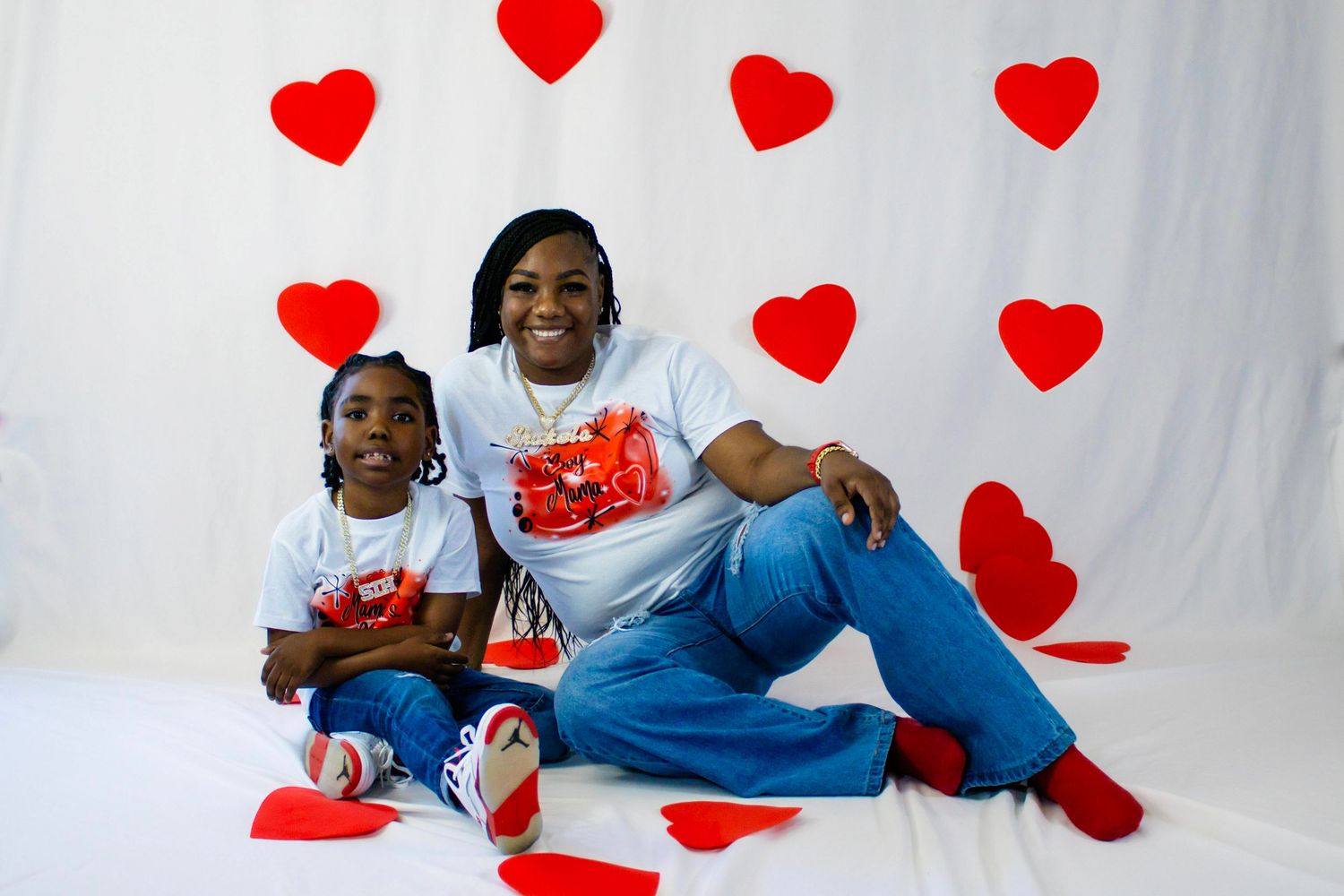 Mother and daughter in matching Valentine's Day shirts sit together in front of heart-decorated backdrop.