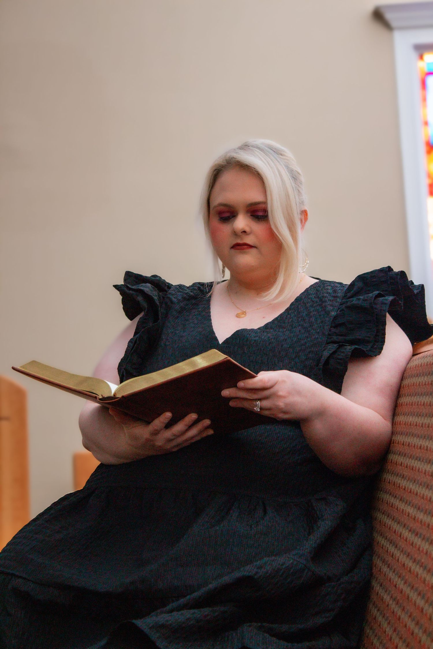 Blonde woman in black dress reading Bible while seated on church pew near stained glass window.