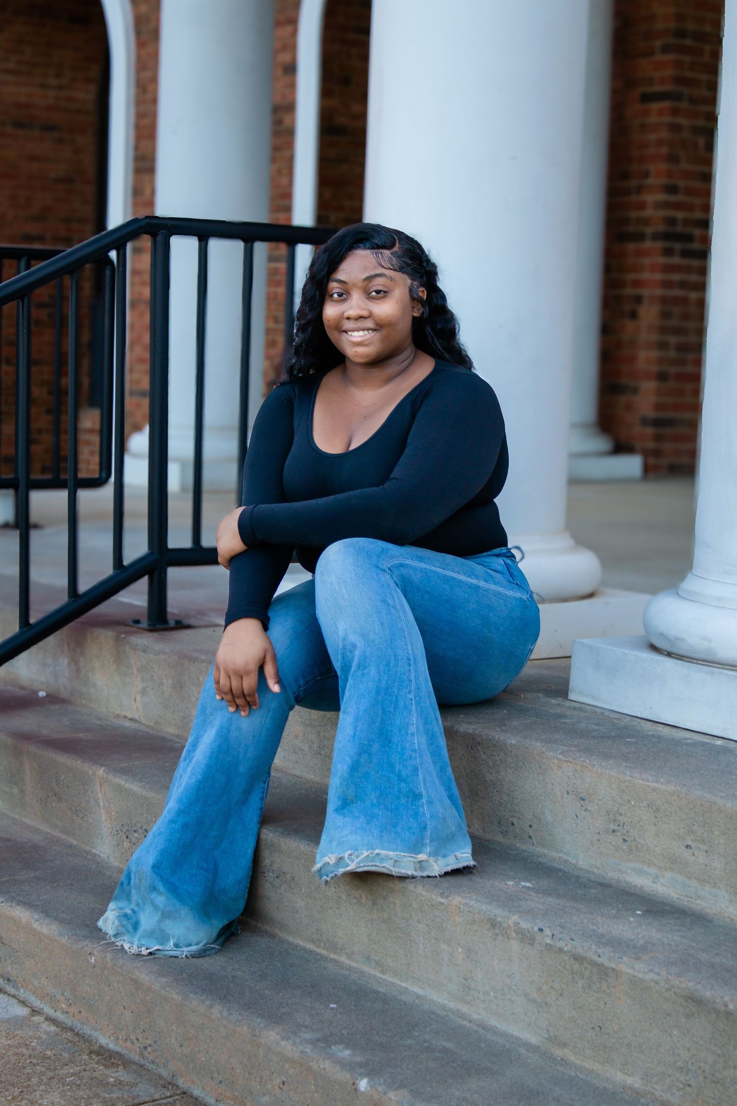 Young Black woman in black long-sleeve top and blue bell-bottom jeans sits on outdoor steps smiling.