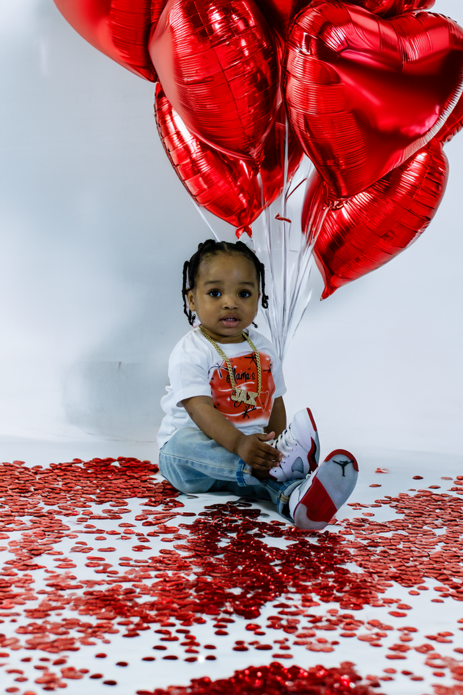 Toddler sitting on floor surrounded by red heart balloons and confetti for Valentine's Day photo.