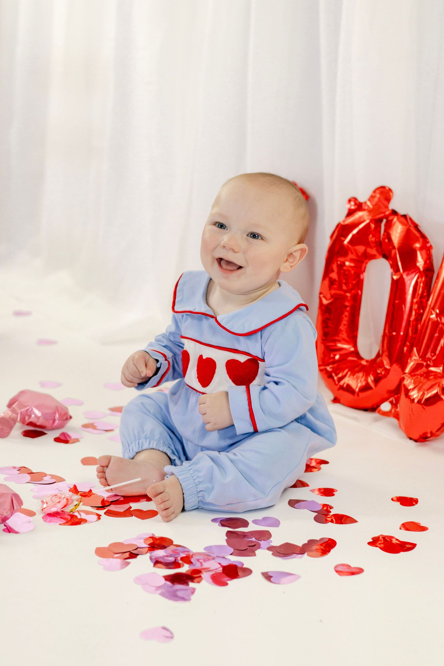 Smiling baby in blue heart romper sits among colorful heart confetti with red balloon decorations for Valentine's Day.