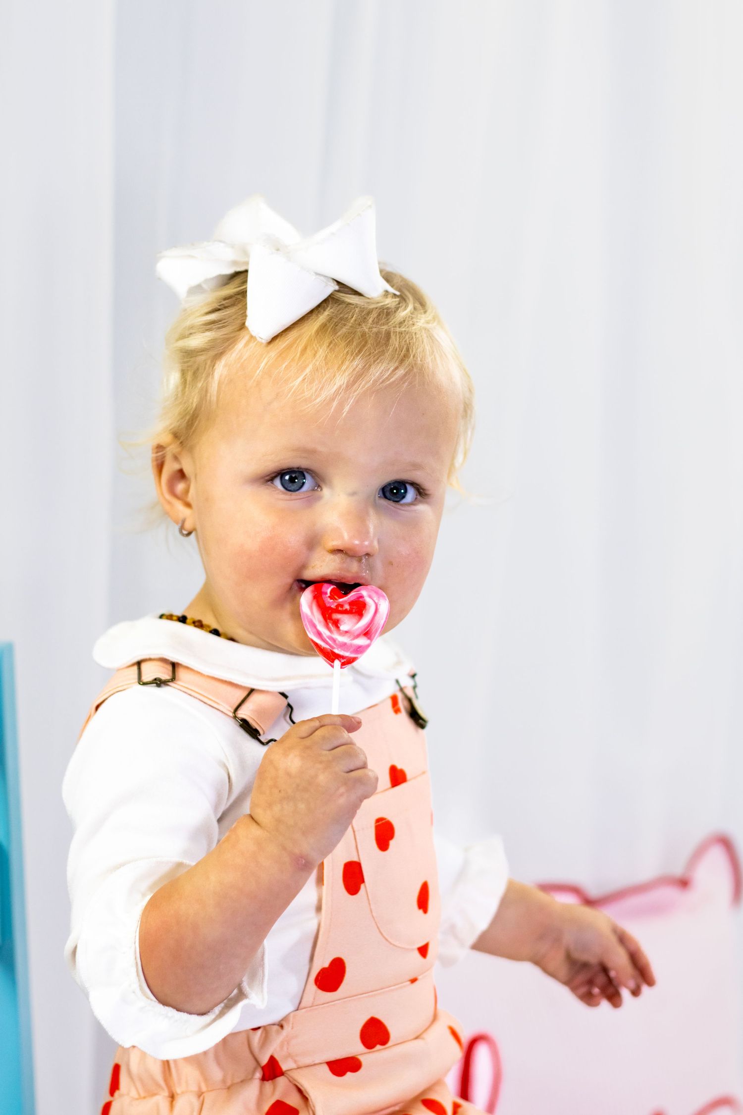 Blonde toddler girl in heart overalls eating a heart-shaped lollipop with white bow in hair.