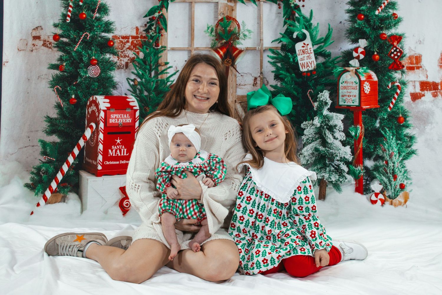 Mother smiles with two daughters in matching Christmas outfits beside decorated holiday trees.