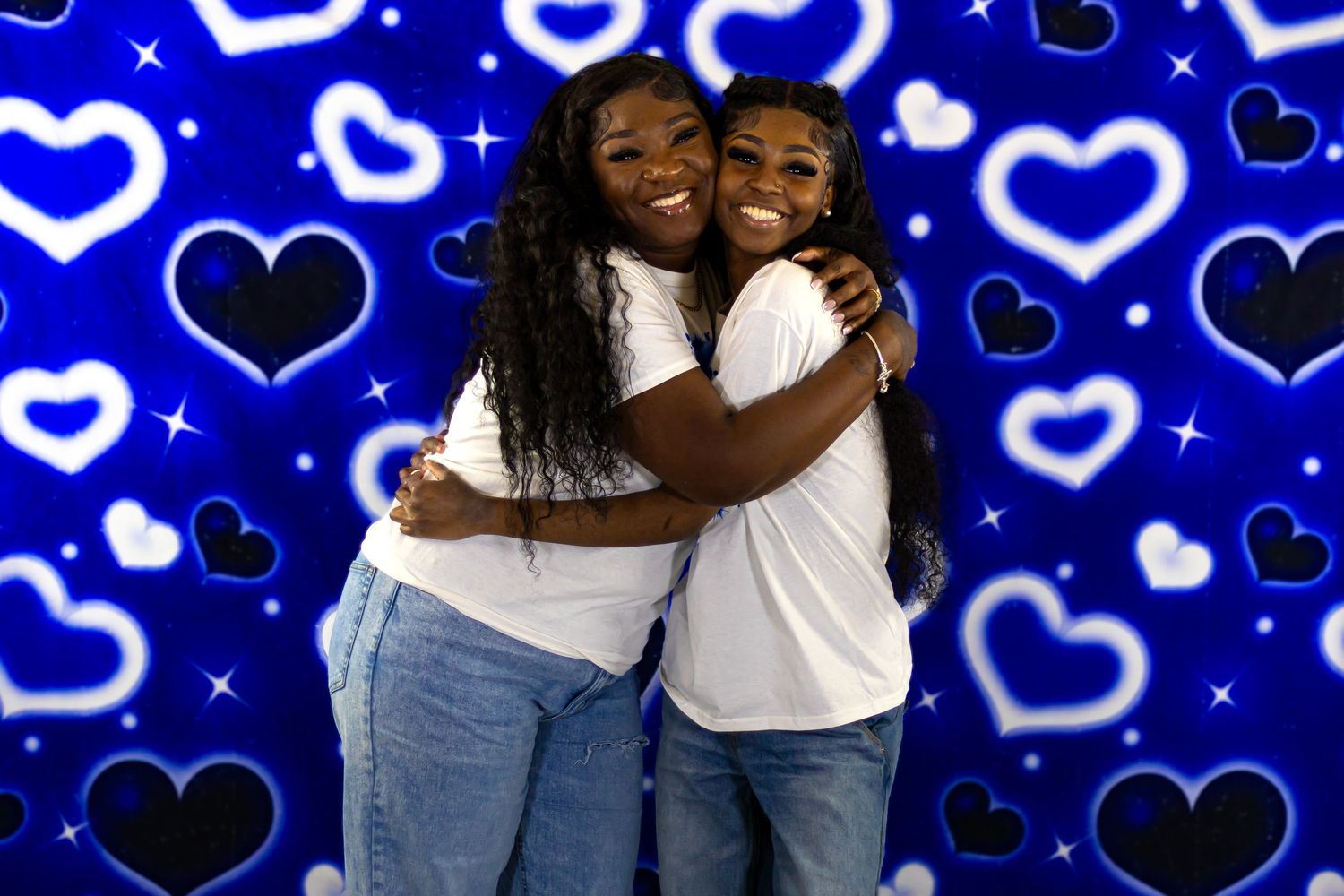 Two women smiling and embracing in white t-shirts against a blue heart backdrop.