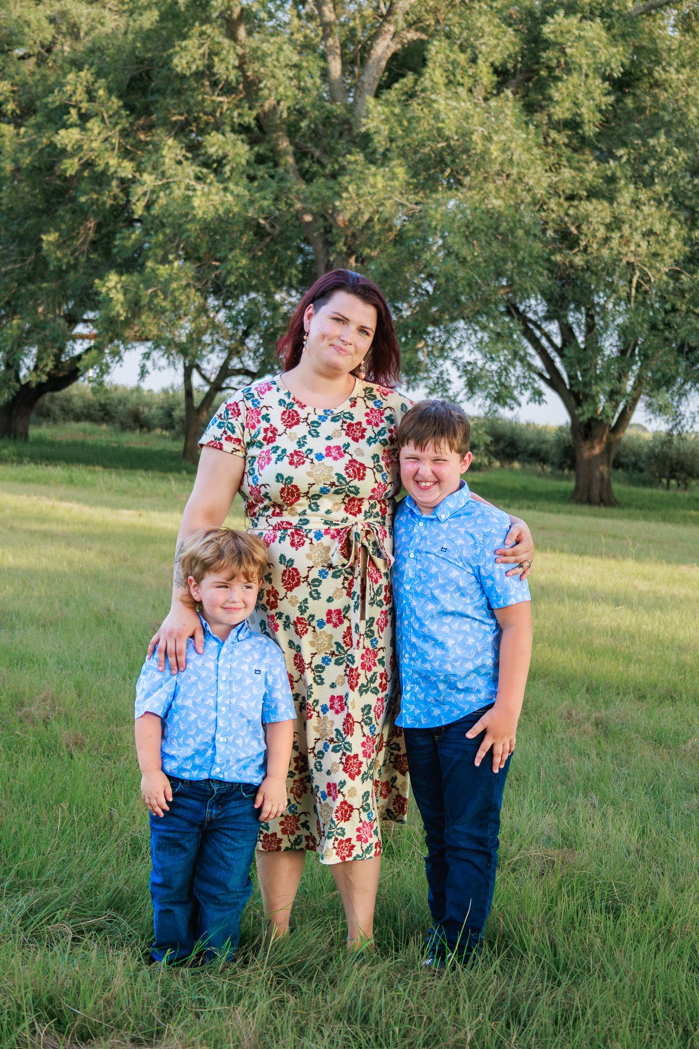 A smiling mother in a floral dress stands with her two boys in blue shirts in a lush green park setting.