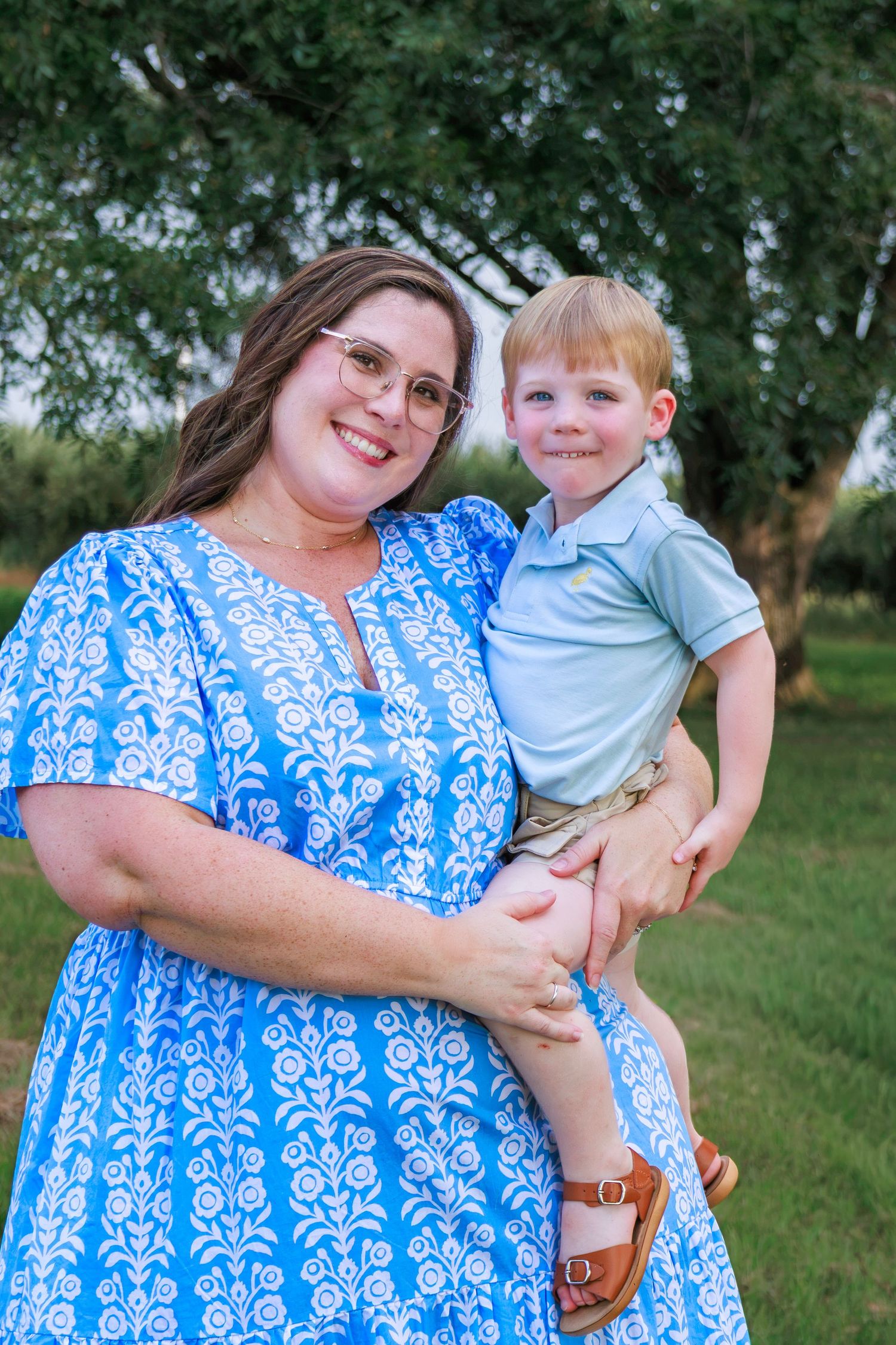 A smiling woman in a blue floral dress holds a young red-haired boy outdoors near green trees.