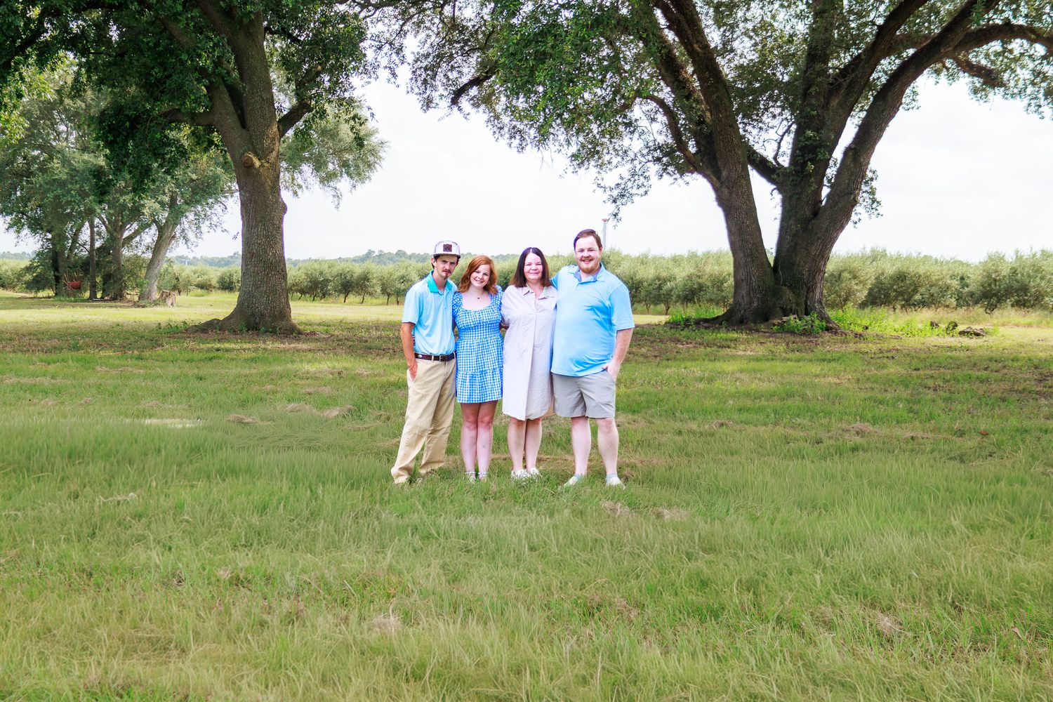 Four family members smiling together on a green lawn with large oak trees in the background.