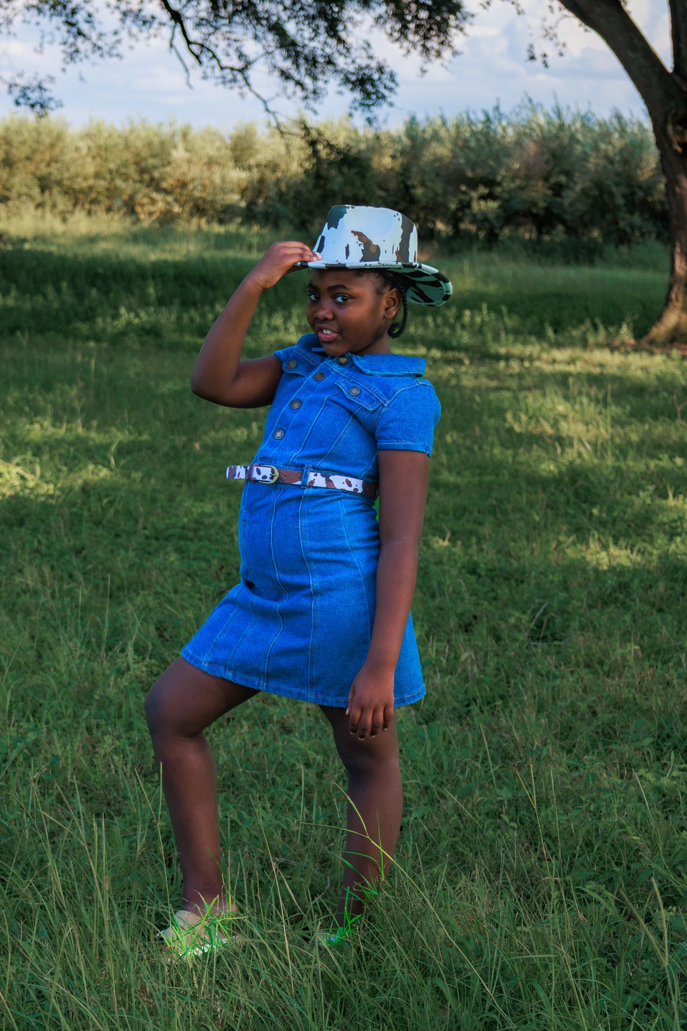 Young girl in blue denim dress and silver hat poses playfully on green grass outdoors.