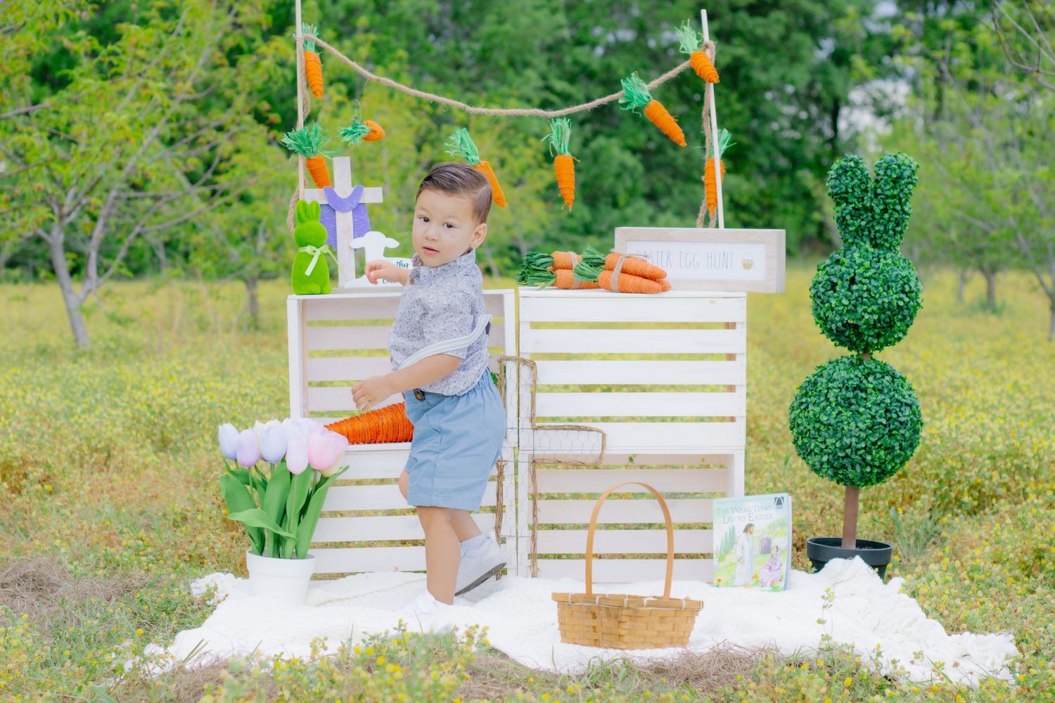 Toddler boy in denim romper standing beside white wooden Easter market stand with carrot decorations outdoors.