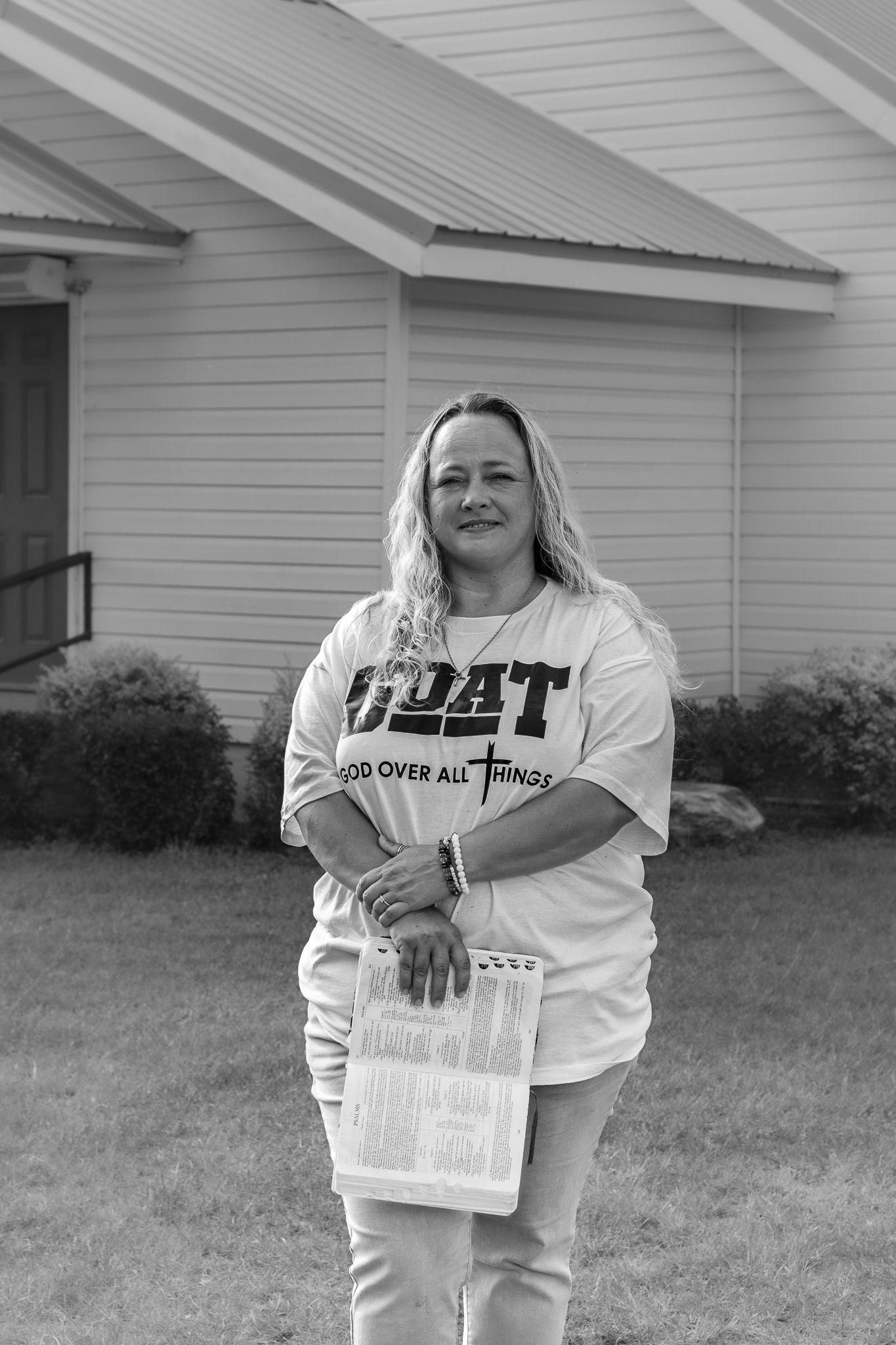 Black and white portrait of woman holding Bible wearing GOAT shirt standing outside white church building.