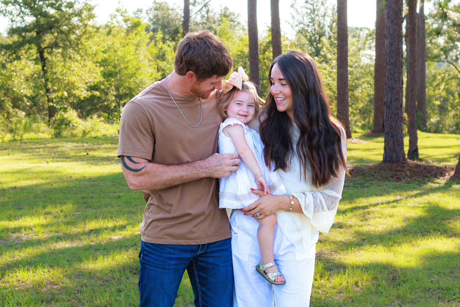 Couple sharing a tender moment with their young daughter in a sunny pine tree park.