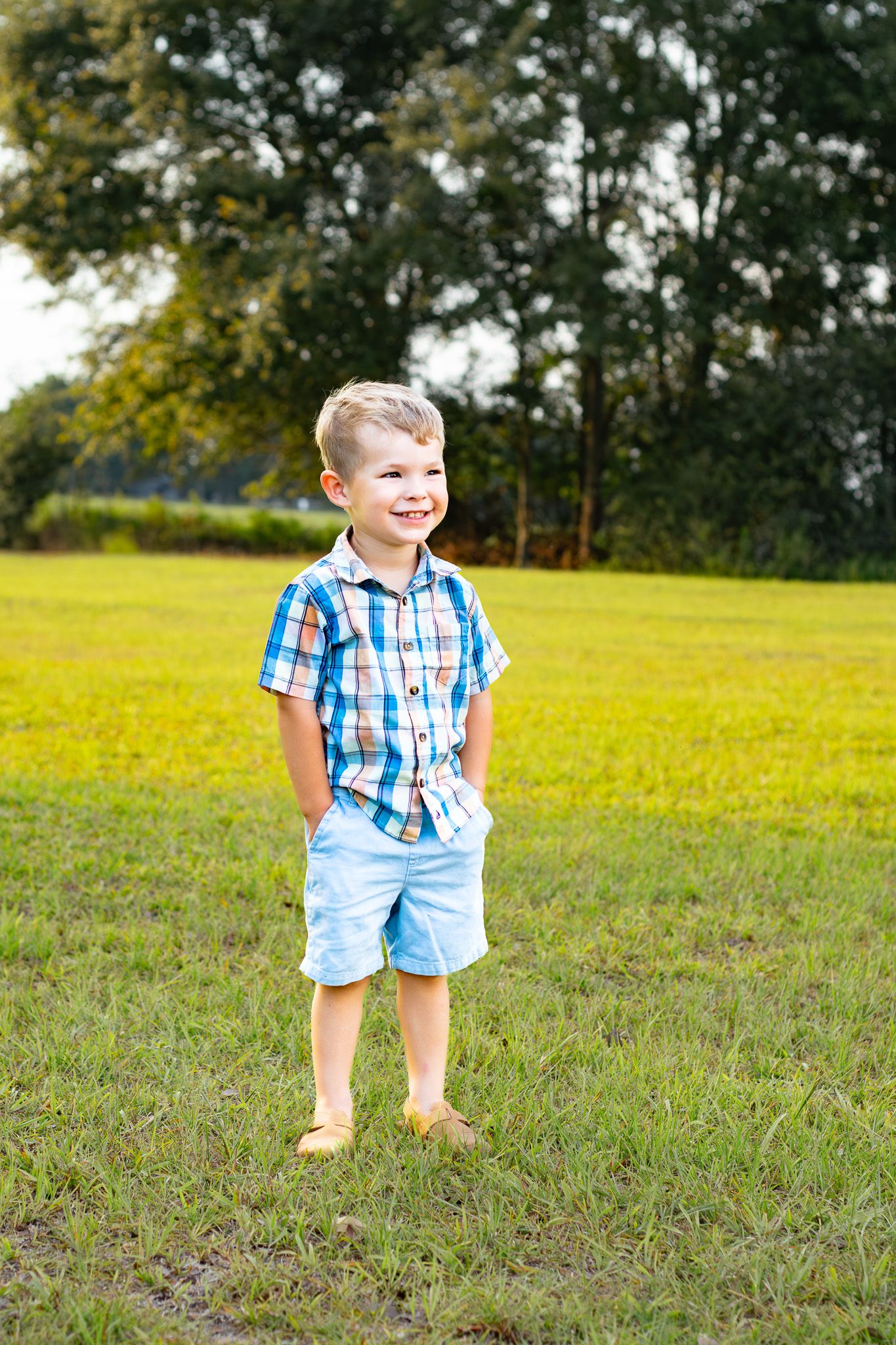 A smiling young boy in a plaid shirt and blue shorts stands barefoot on green grass in a sunny park.